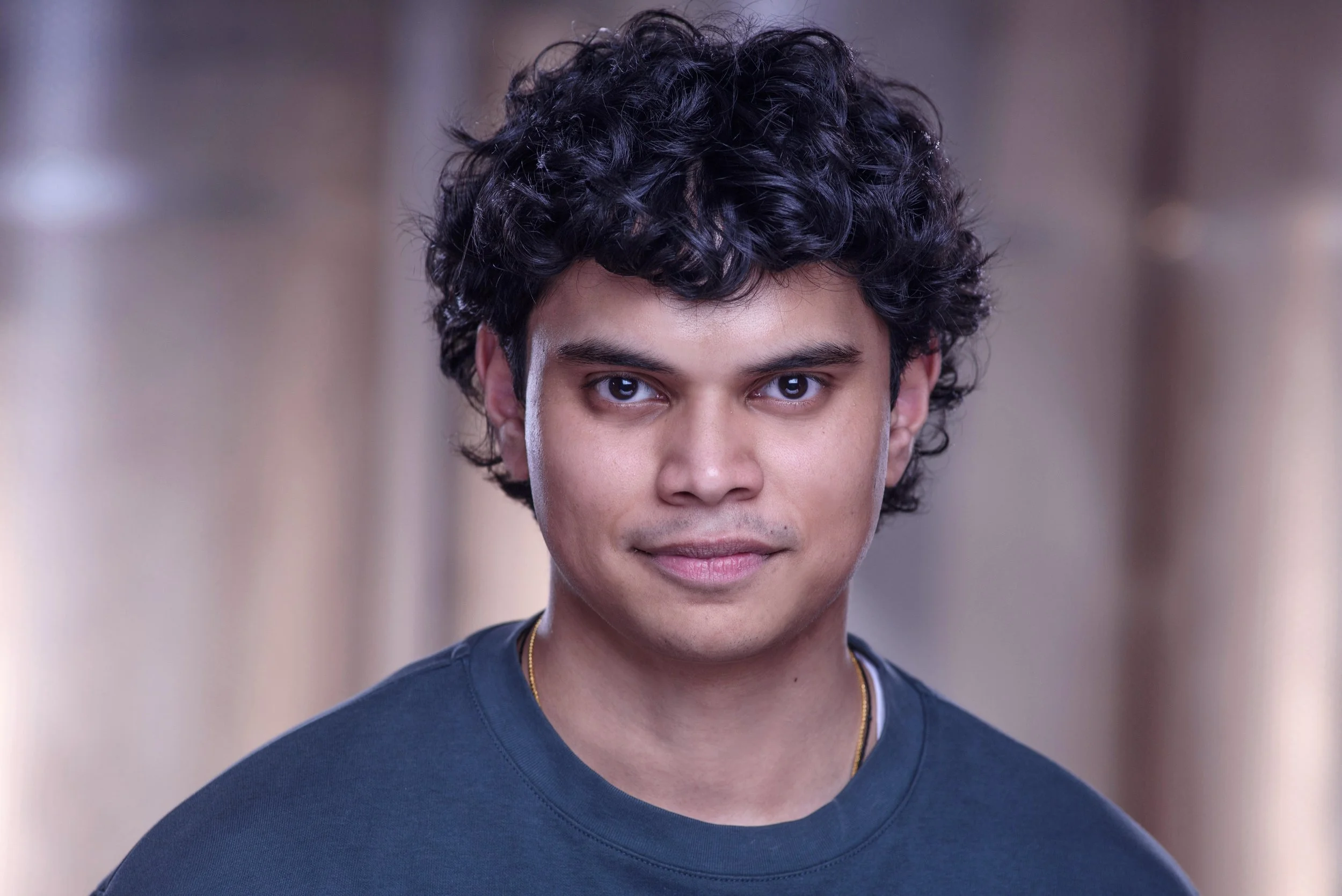 A young man with dark, curly hair and tan skin, wearing a dark blue shirt, looking directly at the camera with a neutral expression.