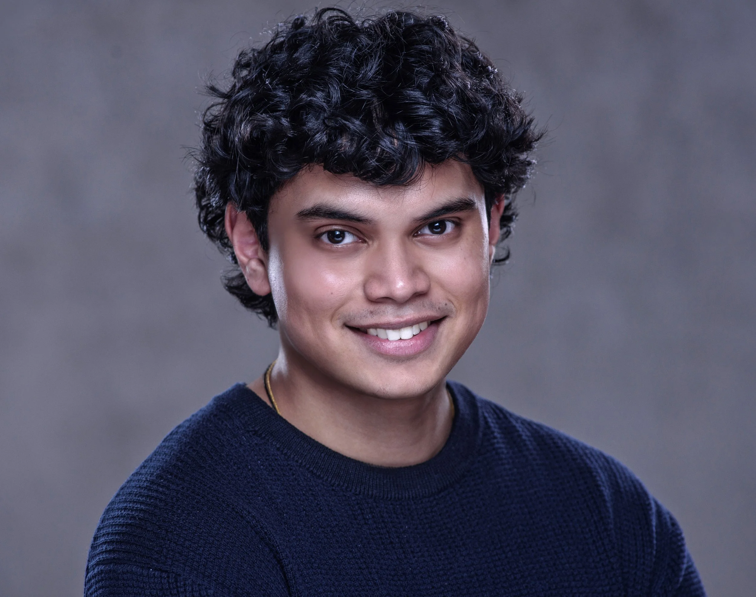 A young man with dark, curly hair smiling and wearing a dark blue sweater against a neutral gray background.