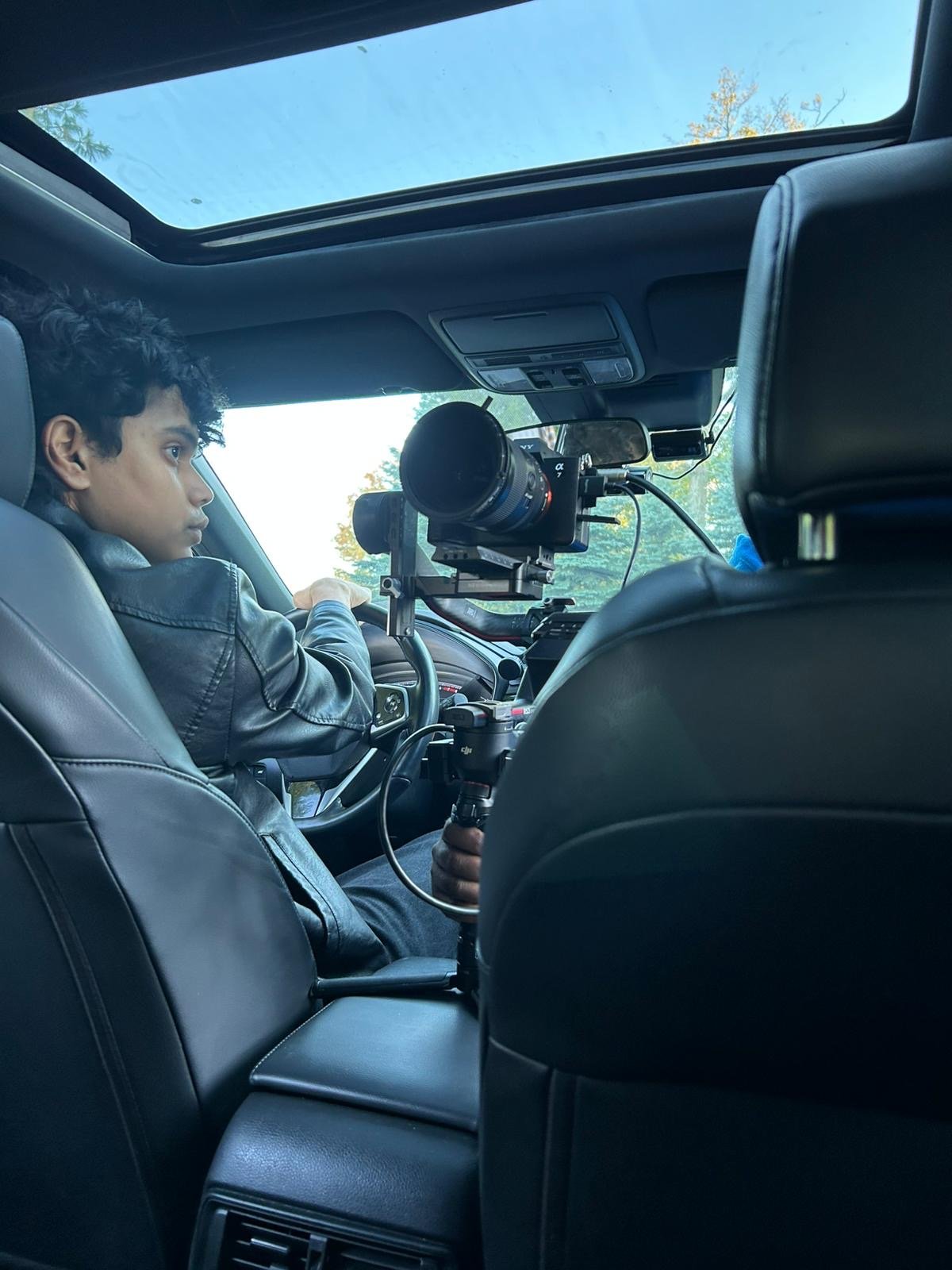 A person with curly hair wearing a leather jacket driving a vehicle with a mounted camera on a stabilizer.