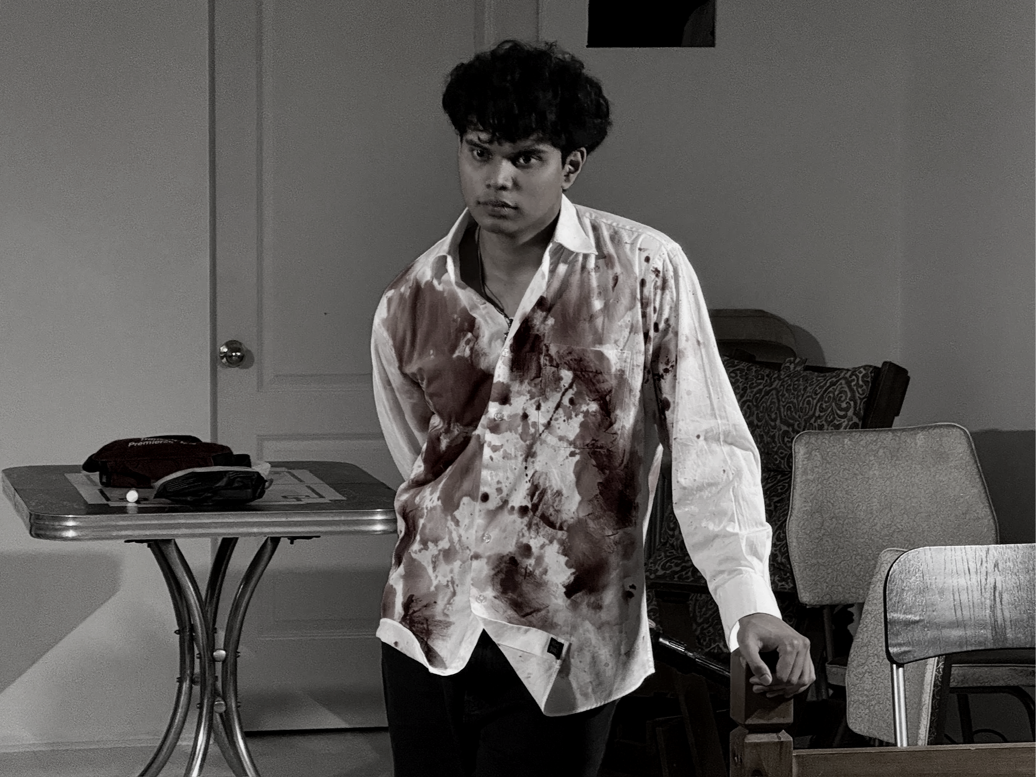 A young man standing in a room wearing a bloodstained white shirt, with dark hair, looking directly at the camera. The room appears to be a casual setting with chairs, a table, and other furniture visible.
