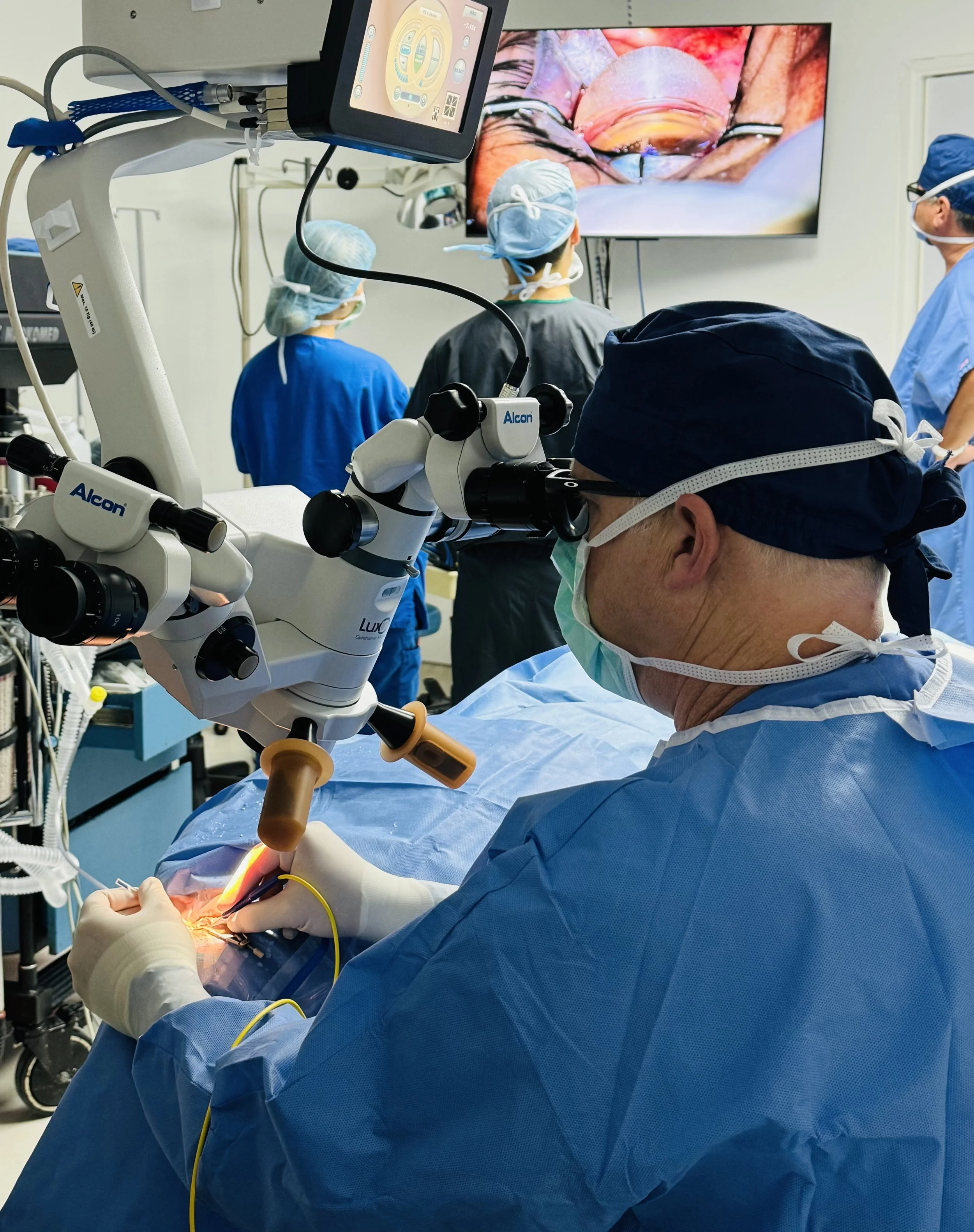 Surgeons performing a surgical operation in an operating room, with a surgeon using a microscope and a monitor displaying a close-up view of the surgery.