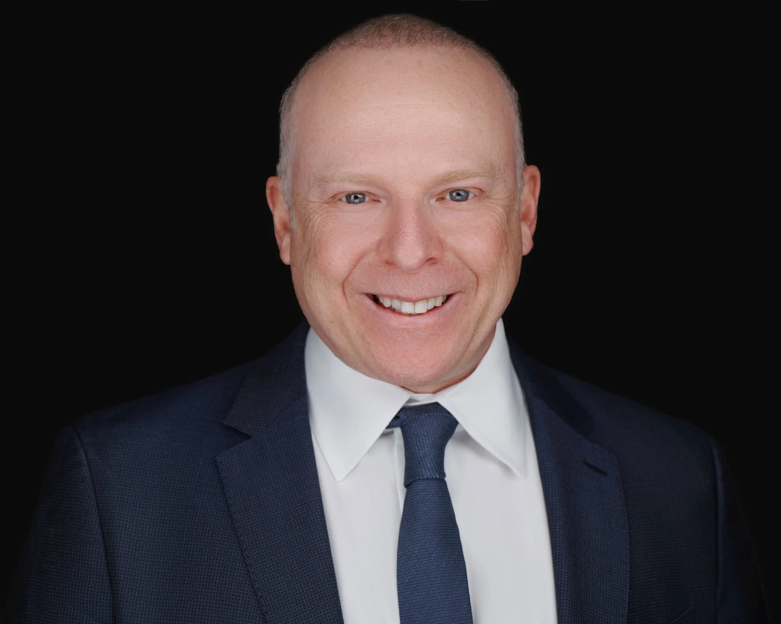 Headshot of a smiling man in a dark suit, white shirt, and blue tie against a black background.
