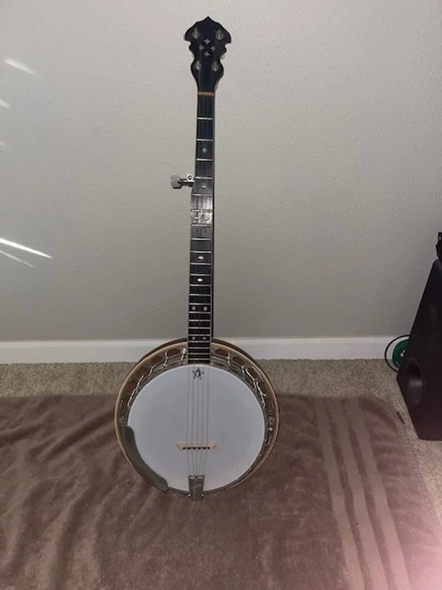 A banjo with a guitar neck attached instead of a traditional body, standing against a plain wall on a carpeted floor.
