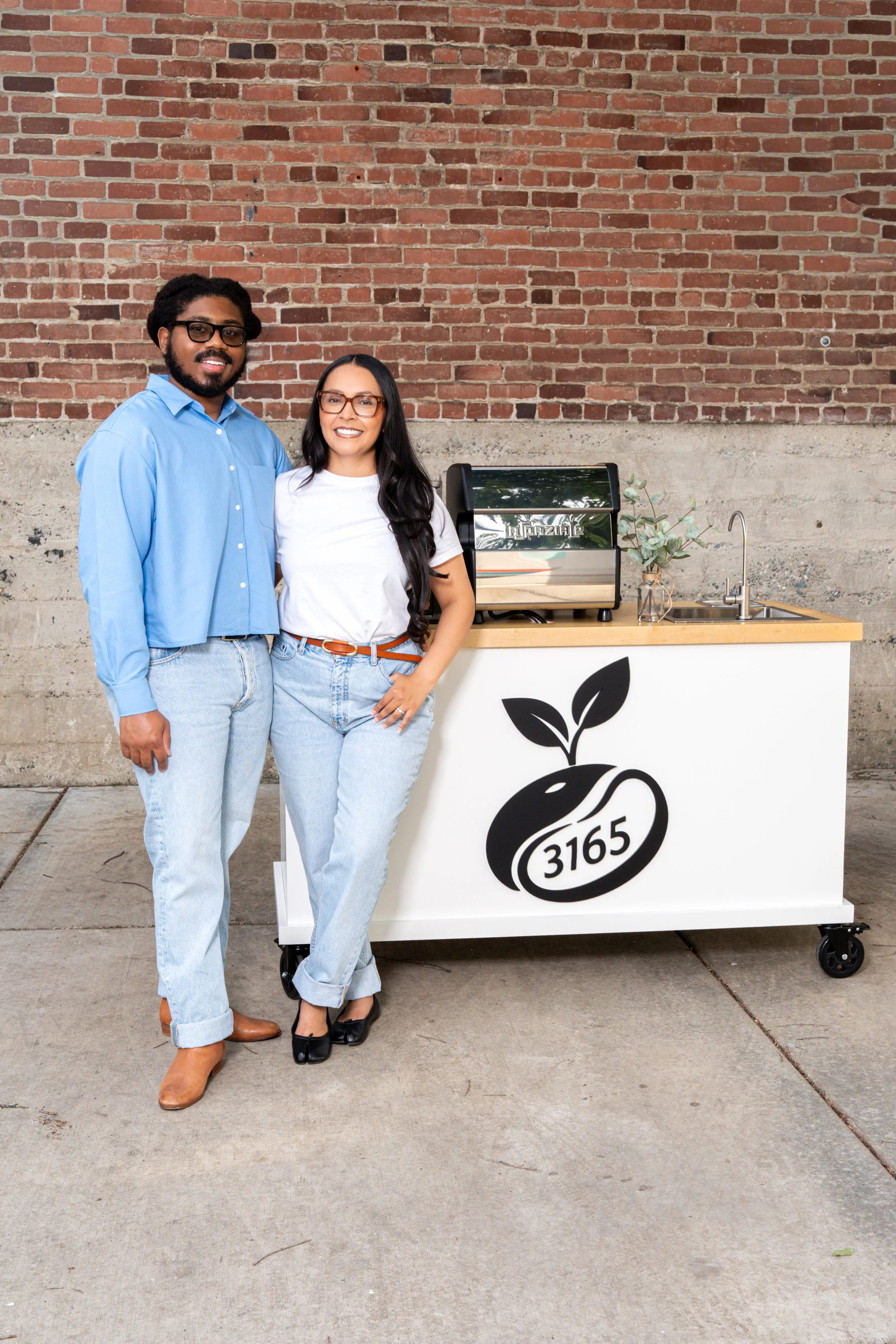 The founders of 3165 Coffee standing beside their modern mobile coffee cart with a custom white espresso bar, eucalyptus decor, and coffee service setup against a red brick wall in Sacramento, California.