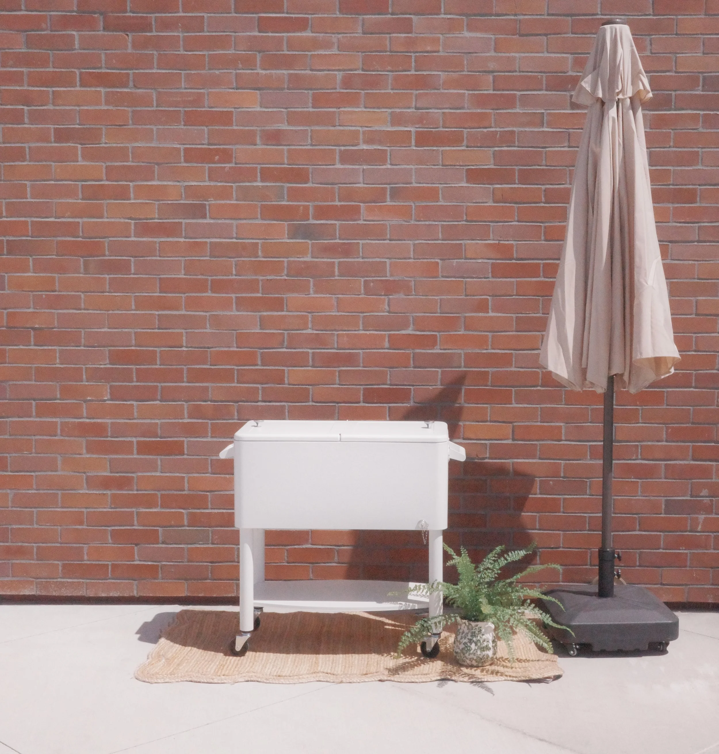 A white beverage cooler with wheels and a beige umbrella displayed beside a potted fern as part of the 3165 Coffee outdoor mobile coffee catering setup against a red brick wall in Sacramento, California.