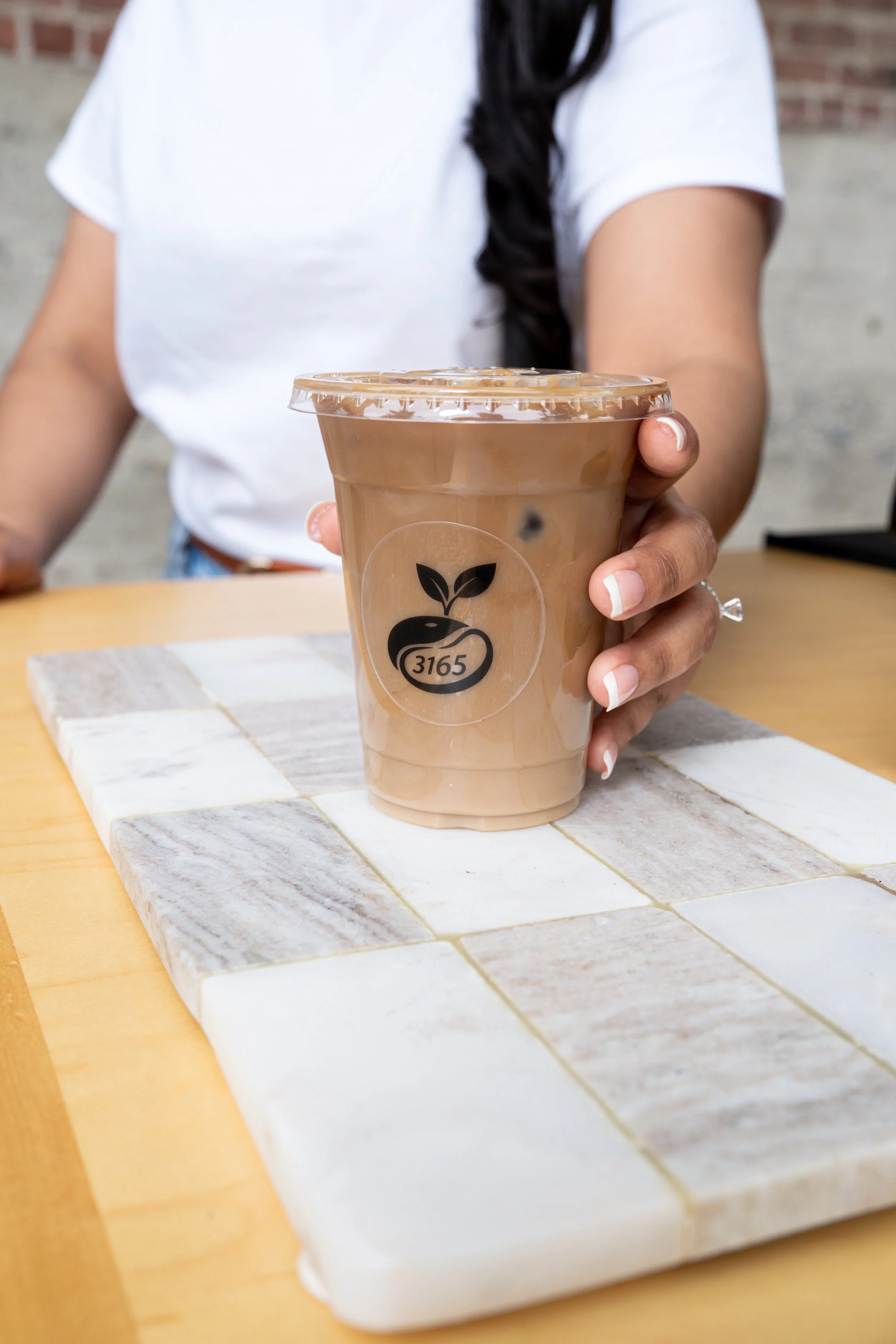 A person holding a handcrafted iced coffee from 3165 Coffee while sitting at a wooden table with beige and white tile detailing in front of a red brick wall in Sacramento, California.