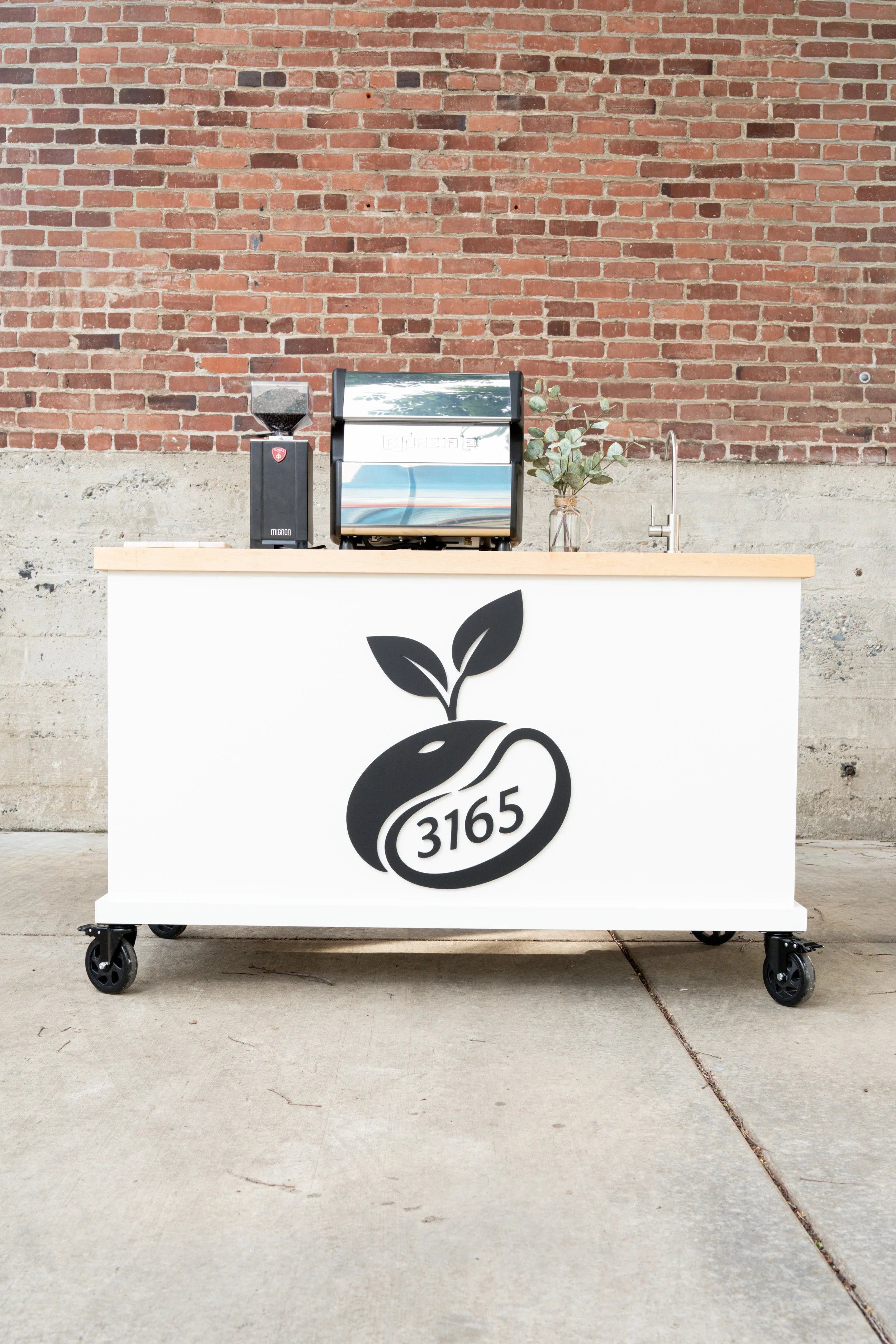 A modern 3165 Coffee mobile espresso cart with a white front panel, black coffee bean logo, wooden countertop, espresso machine, coffee grinder, and fresh eucalyptus decor set up against a red brick wall in Sacramento, California.
