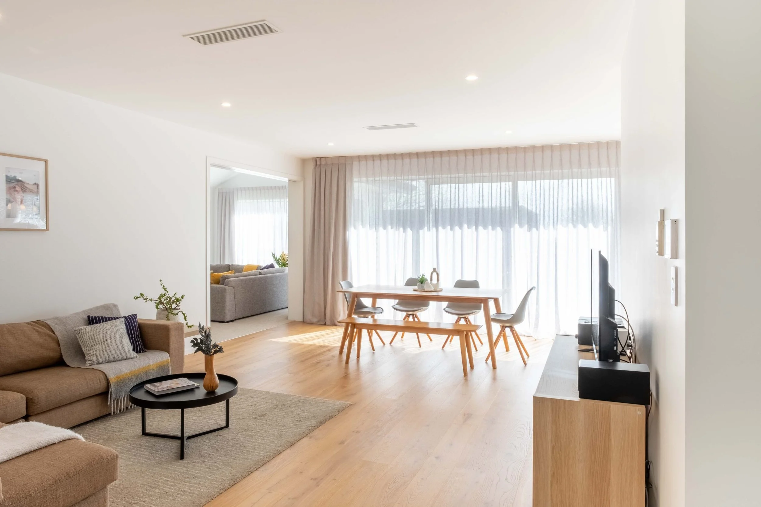 Bright living and dining room with large window, beige sofa, coffee table, dining table with chairs, and TV.