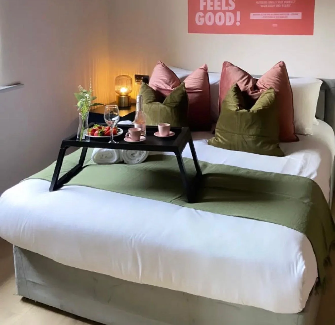 A bed with a green and white bedspread, topped with a black tray holding a glass bottle of water, a wine glass, pink cups, a small plate of strawberries, and a plant. There are pink and green pillows on the bed, and a small bedside table with a lit l