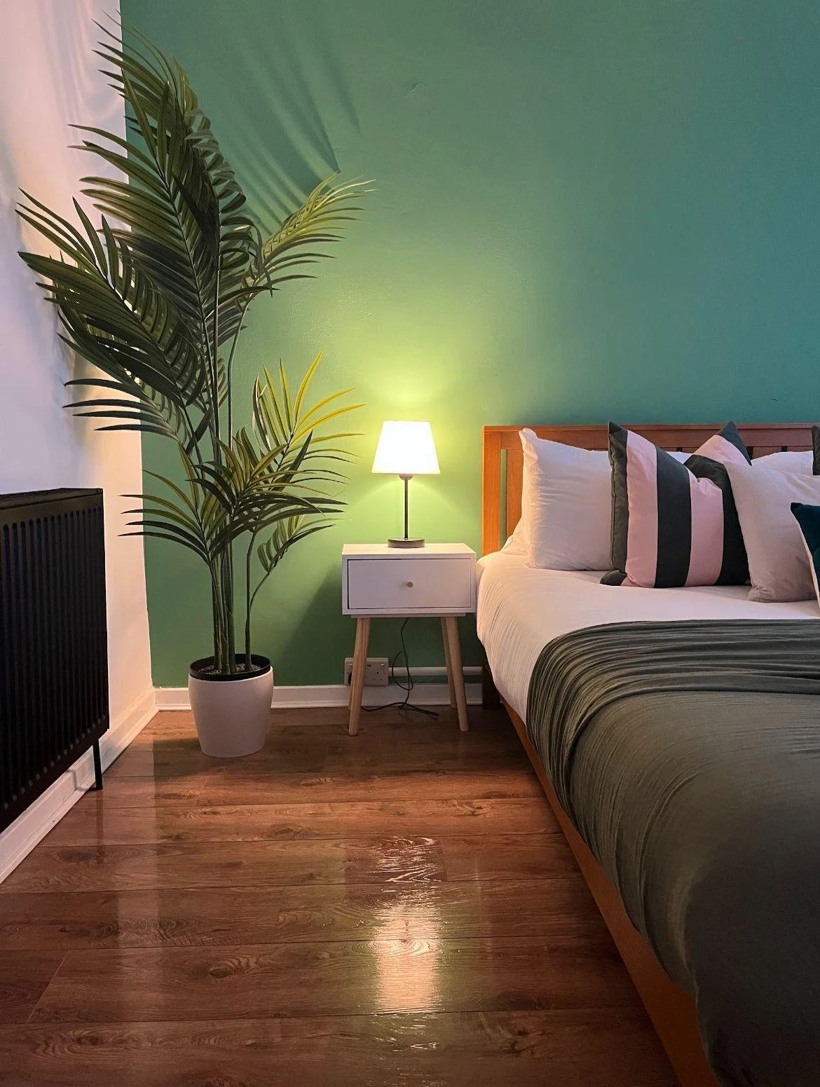 A bedroom with a green accent wall, a wooden bed with white and dark pillows, a white nightstand with a lamp, a potted plant, and hardwood flooring.