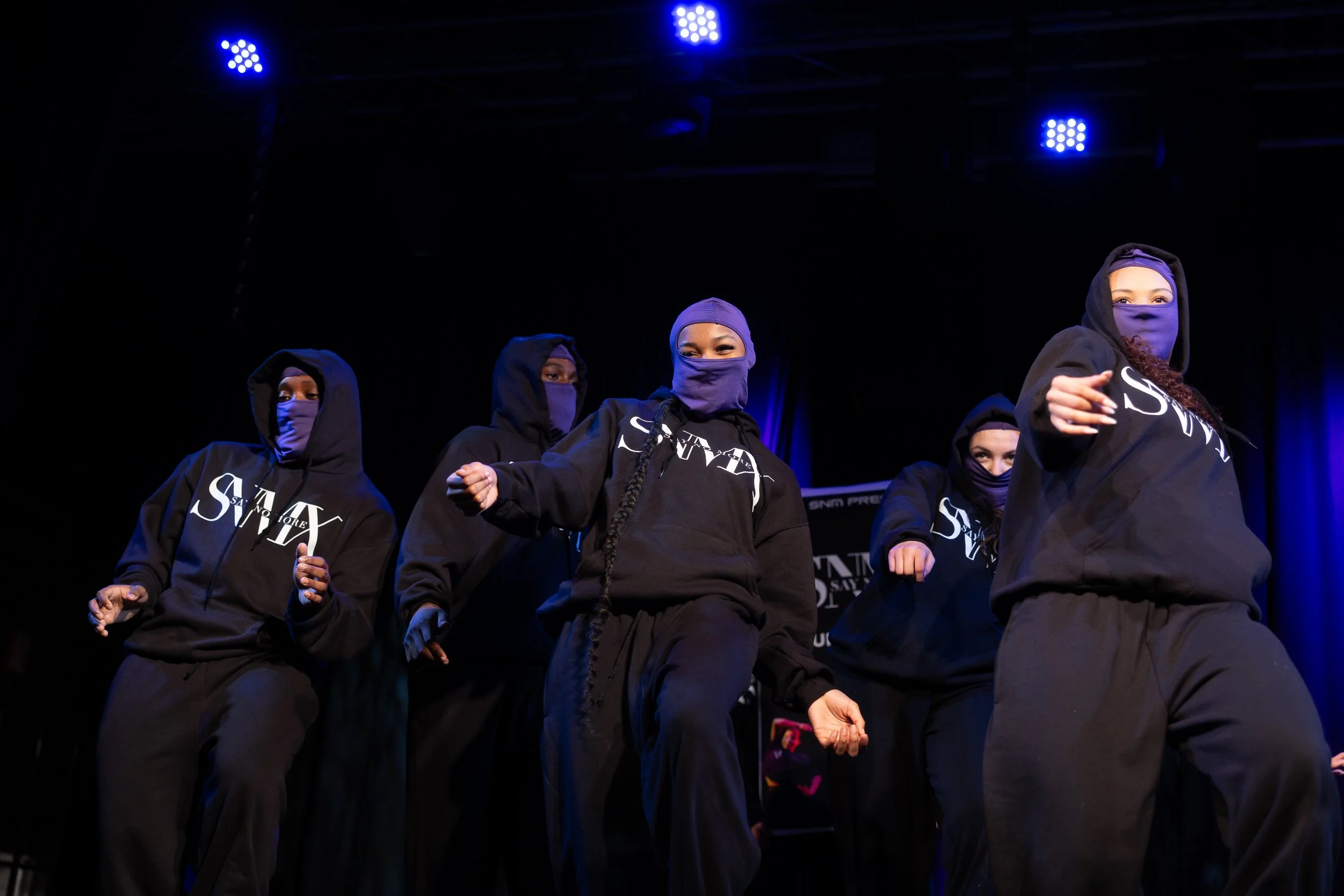 A group of diverse women wearing black hoodies and masks with the word 'SAVIOR' printed in white, performing a dance routine on a stage illuminated by blue lights.
