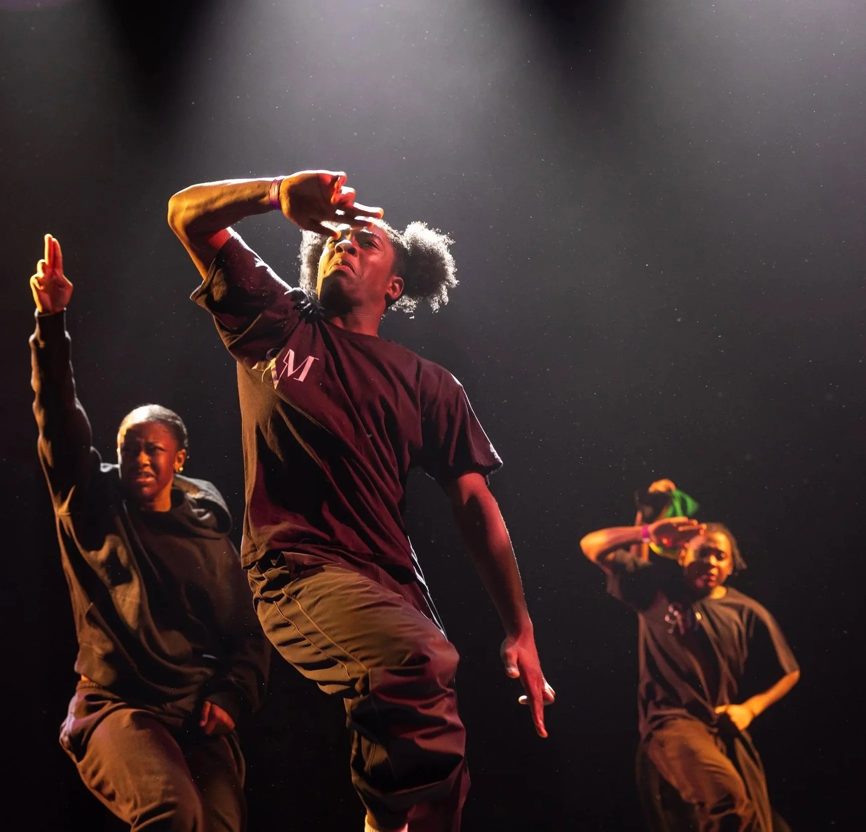 A group of young dancers performing on a stage, with one dancer prominently in the center making a gesture with his hand and looking serious, under stage lights.