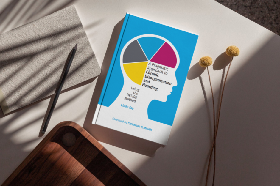 Book titled 'A Pragmatic Approach to Chronic Disorganisation and Hoarding' by Linda Fay on a white surface, accompanied by a pen, a notepad, and dried yellow flowers.