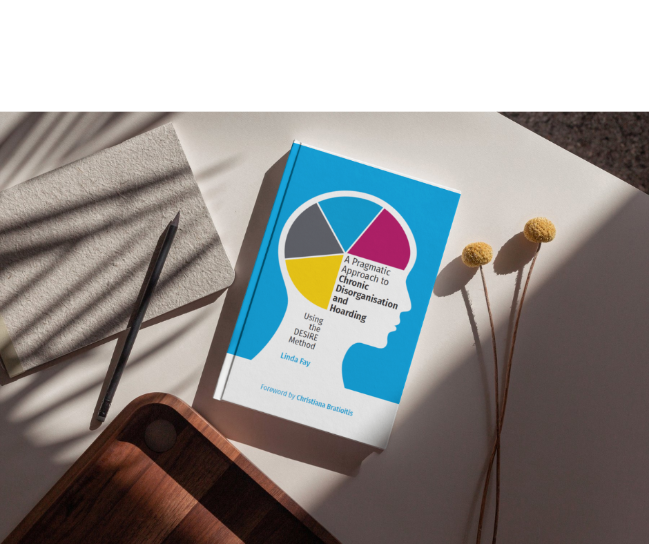 Book titled 'A Pragmatic Approach to Chronic Disorganisation and Hoarding' by Linda Fay, with foreword by Christina Bratiotis, on a white surface with two yellow billy balls, a gray notepad, a black pen, and part of a wooden tray and shadowed plant leaves.