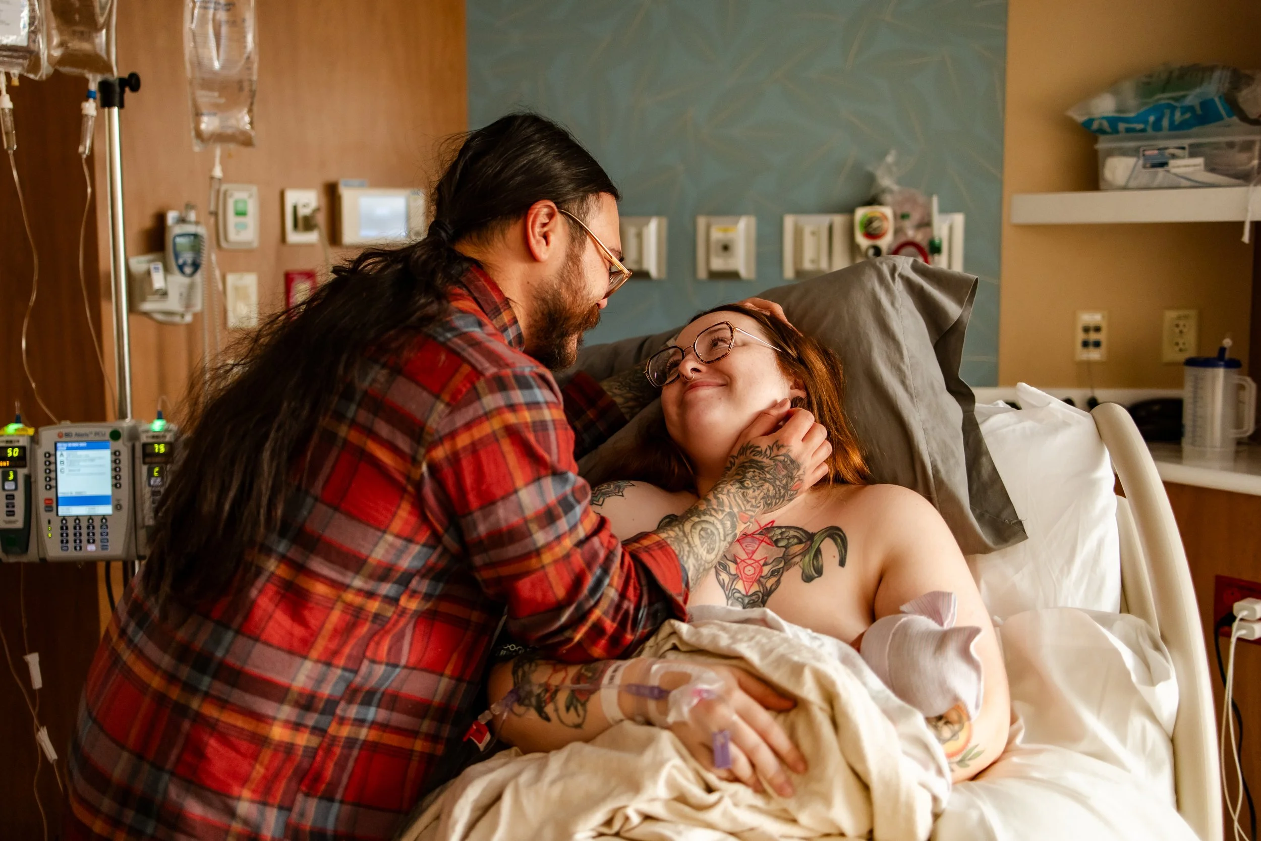 A man tenderly holds a woman's face as he leans over her in a hospital bed, smiling. The woman is lying in a hospital bed, with tattoos on her arm and chest, and productivity IV lines. Both appear happy and connected.