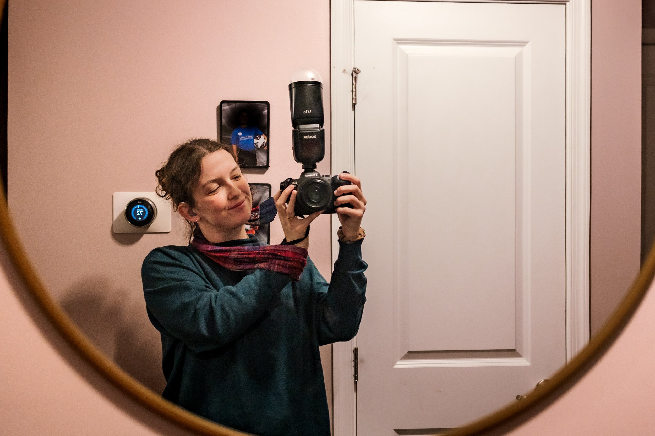 A woman taking a selfie in a circular mirror with her camera. She is smiling with eyes closed, wearing a dark jacket and a red scarf. The background shows a pink wall, a thermostat, a white door, and a small photo frame.