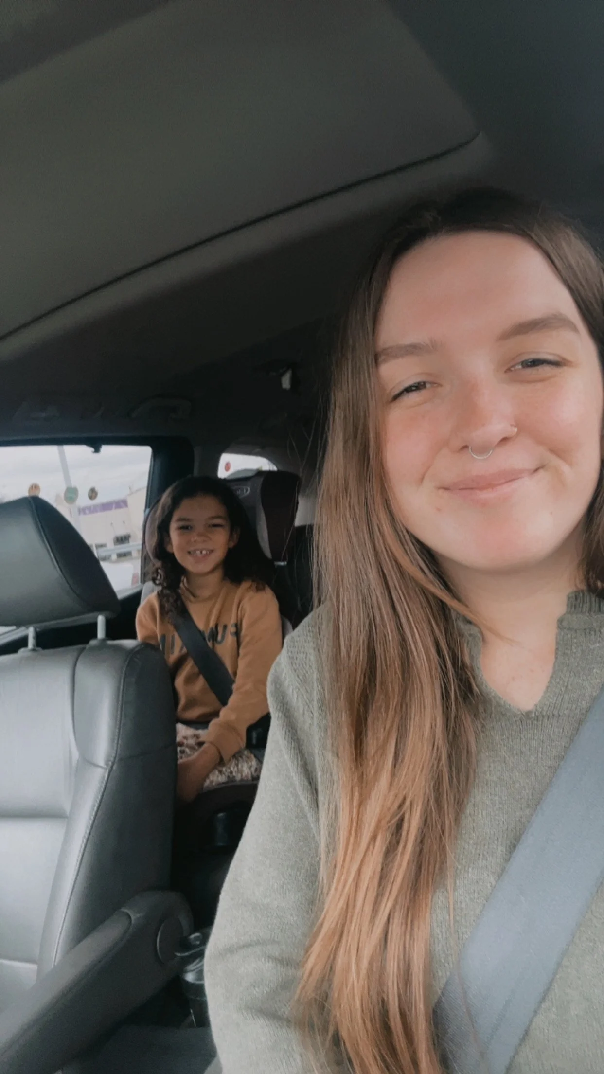 A woman with long brown hair and a septum piercing taking a selfie in a car, with a young girl sitting in the back seat smiling, both wearing seatbelts.