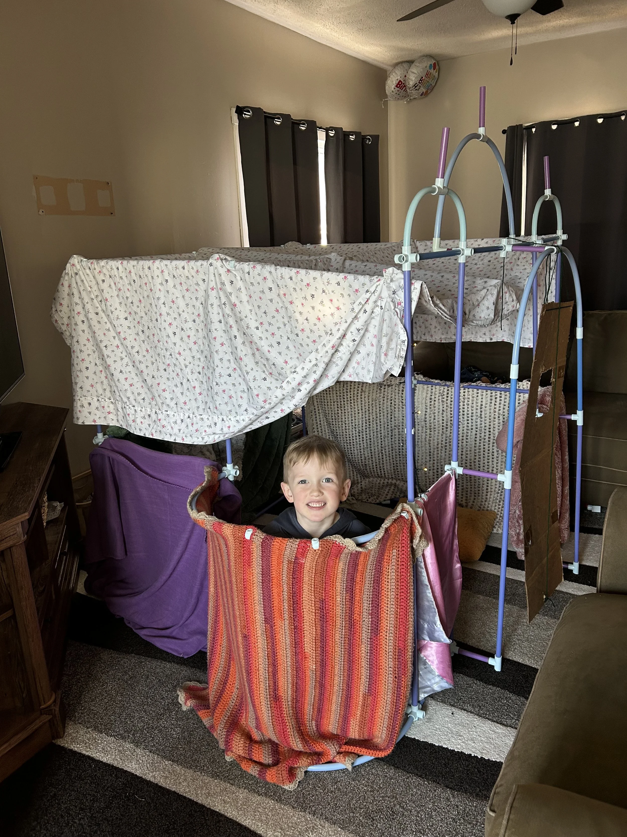 A young boy smiling inside a makeshift indoor play structure made of bed sheets, blankets, and a bed canopy, set up in a living room.