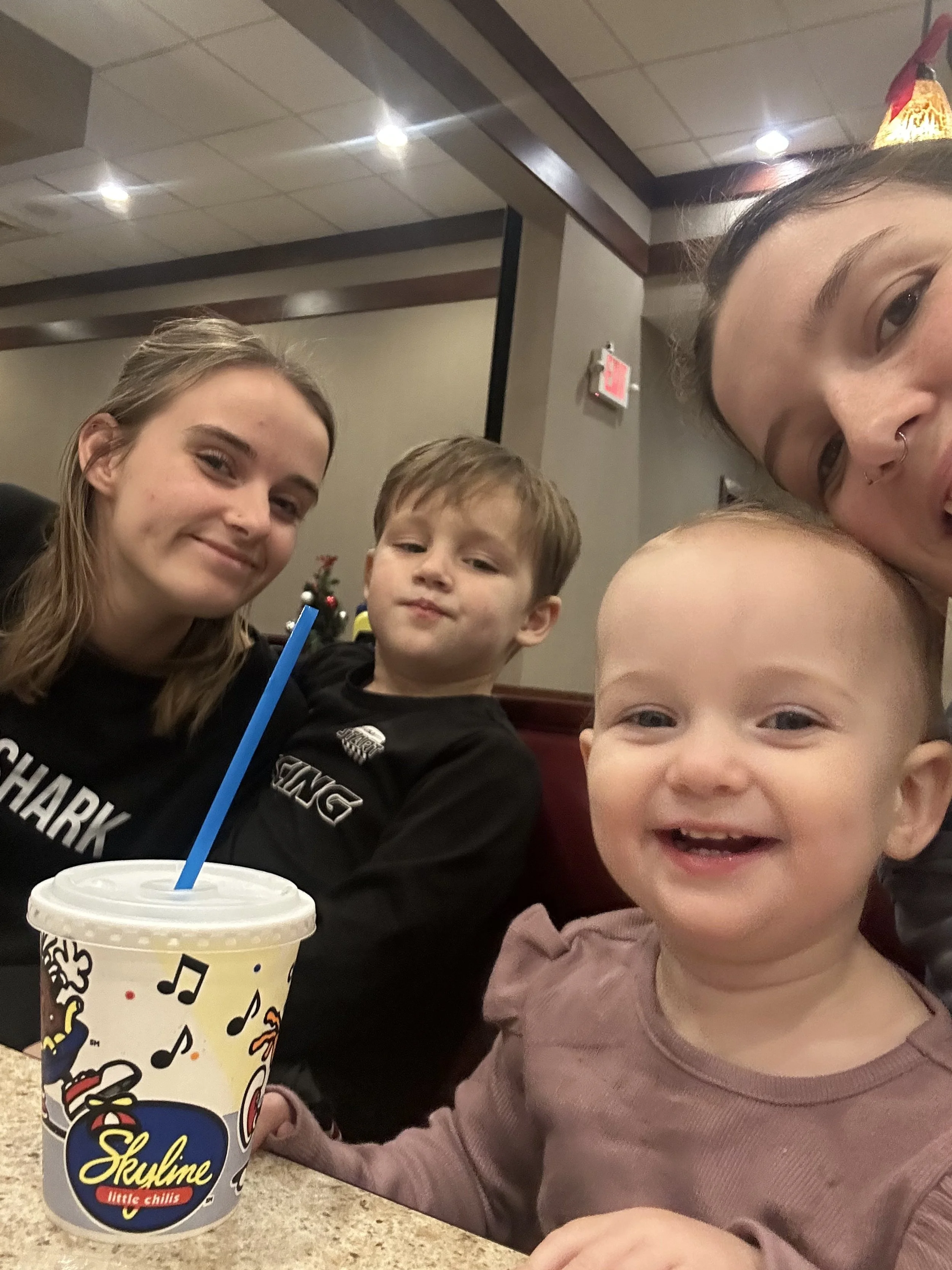 A group of four people, two women and two children, sitting at a table in a restaurant. They are smiling; the woman on the right is partially visible. A large cup with a blue straw and colorful music notes and cartoon characters is on the table in fr
