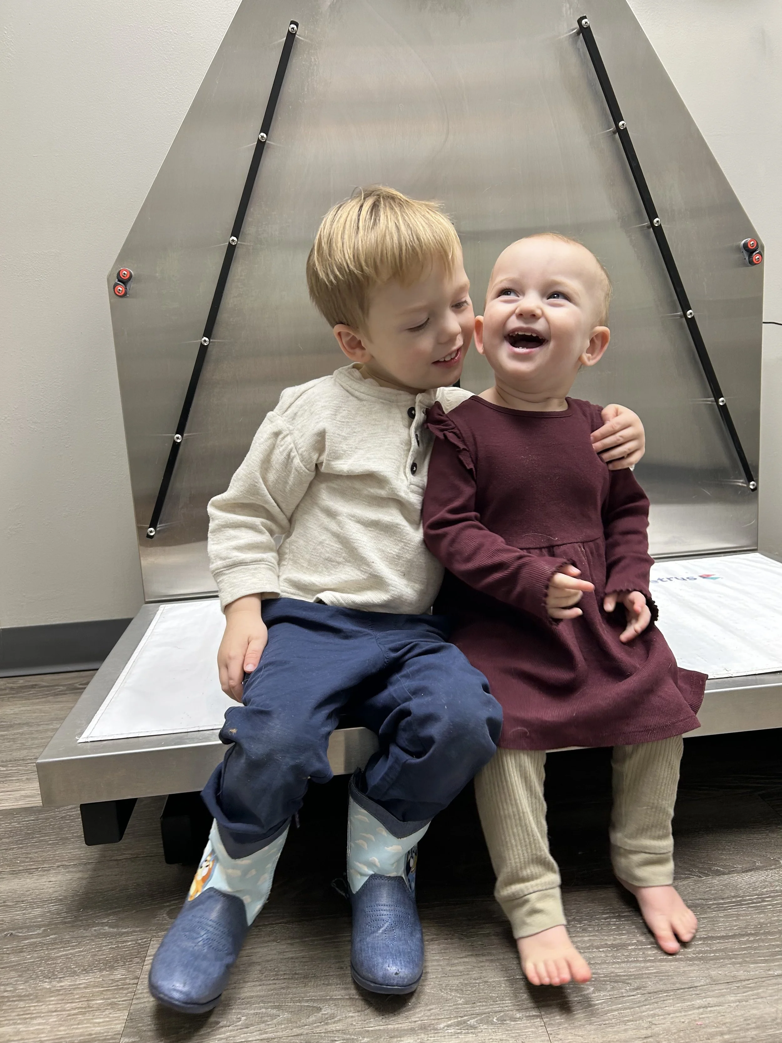 Two young children, a boy and a girl, are sitting close together and smiling. The boy has light brown hair and is wearing a beige shirt, navy pants, and rain boots. The girl has light hair and is wearing a burgundy dress and beige leggings. They are 