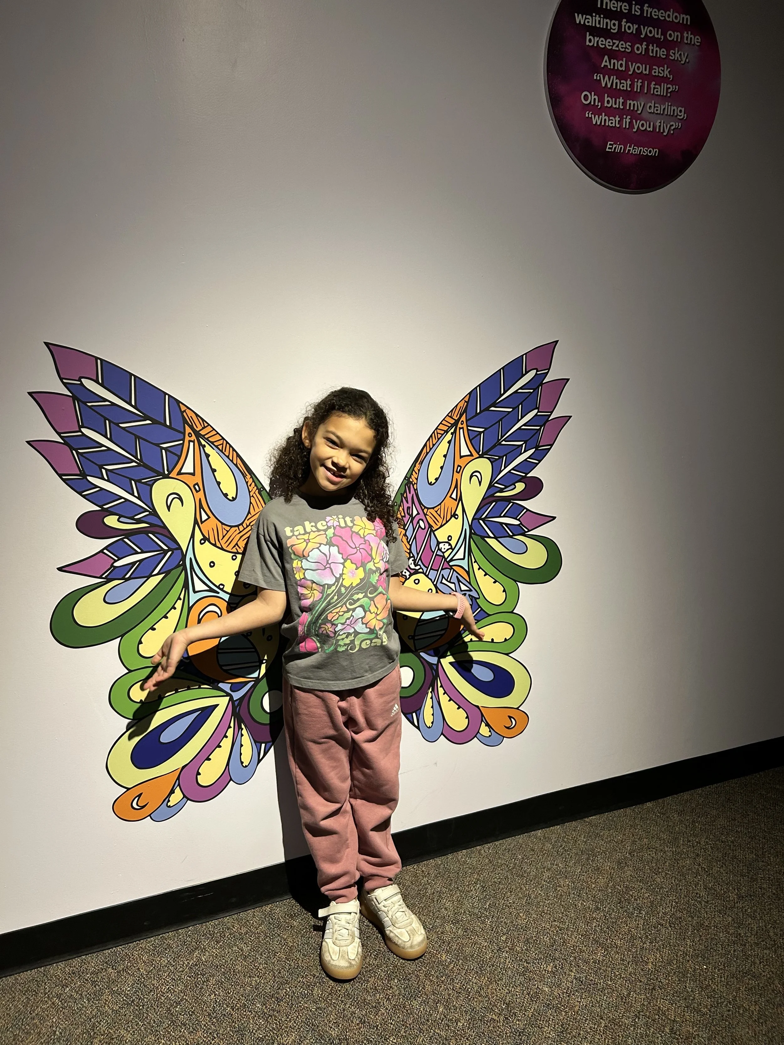 A young girl standing in front of wall with a large, colorful butterfly wings mural, smiling and posing with arms outstretched.
