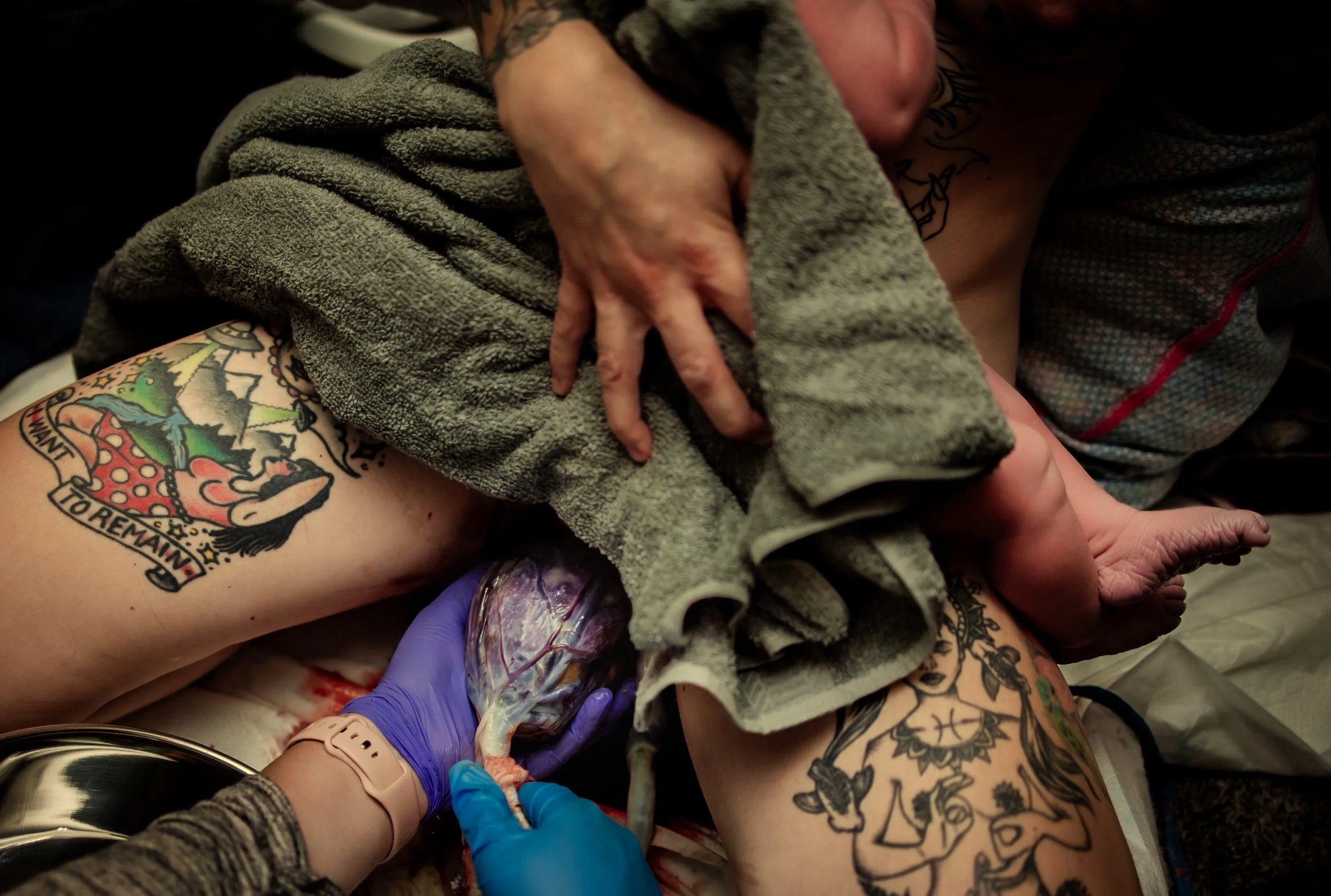 A person giving birth, with a baby emerging from their body while another person assists and supports, holding the baby with gloves, and tattoos are visible on the person's arm.