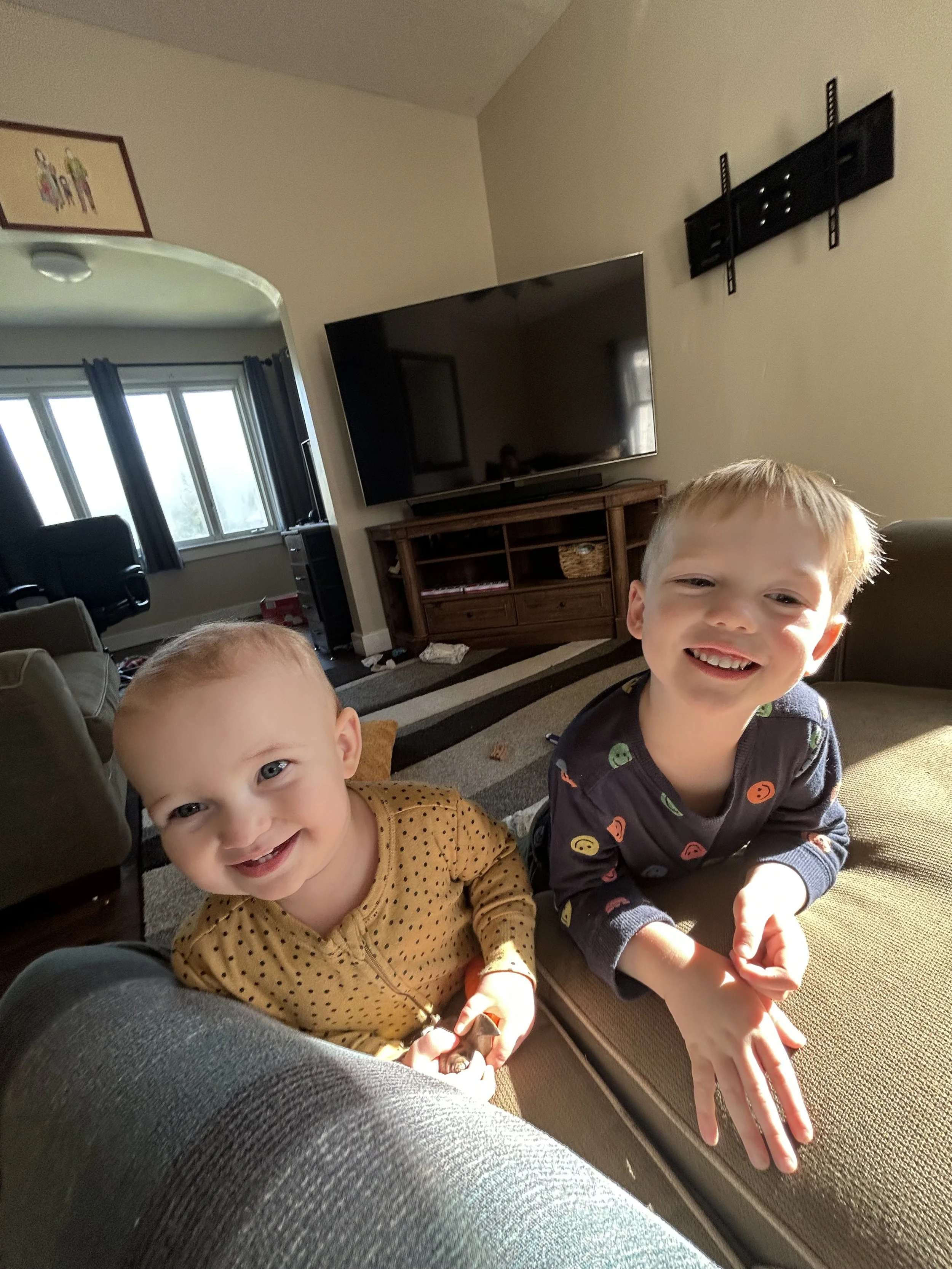 Two young children smiling and leaning over the back of a couch in a living room. The background features a large television mounted on the wall, a wooden TV stand, and a window with dark curtains.