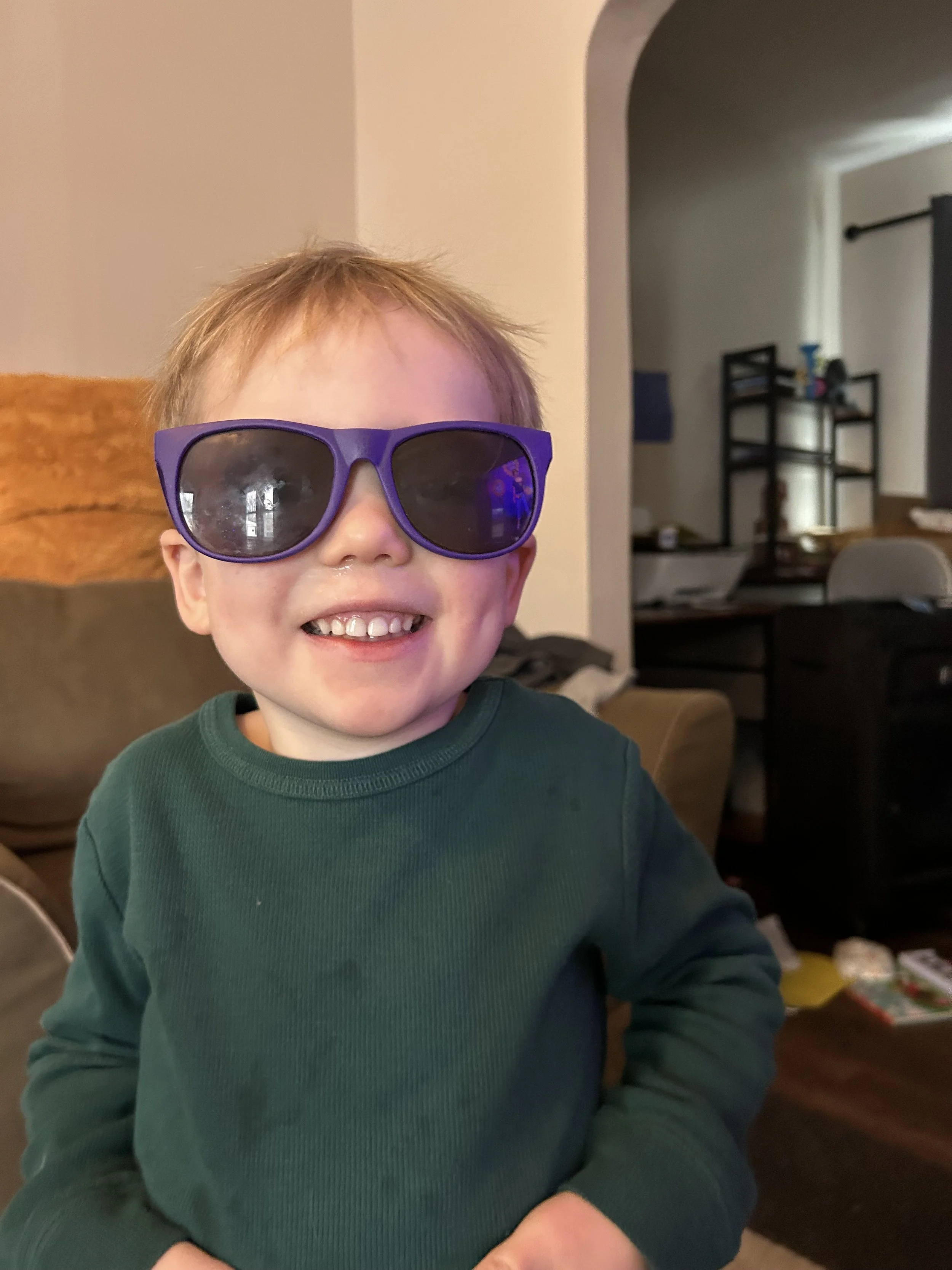 A young boy with light brown hair smiling, wearing large purple sunglasses and a green long-sleeve shirt.