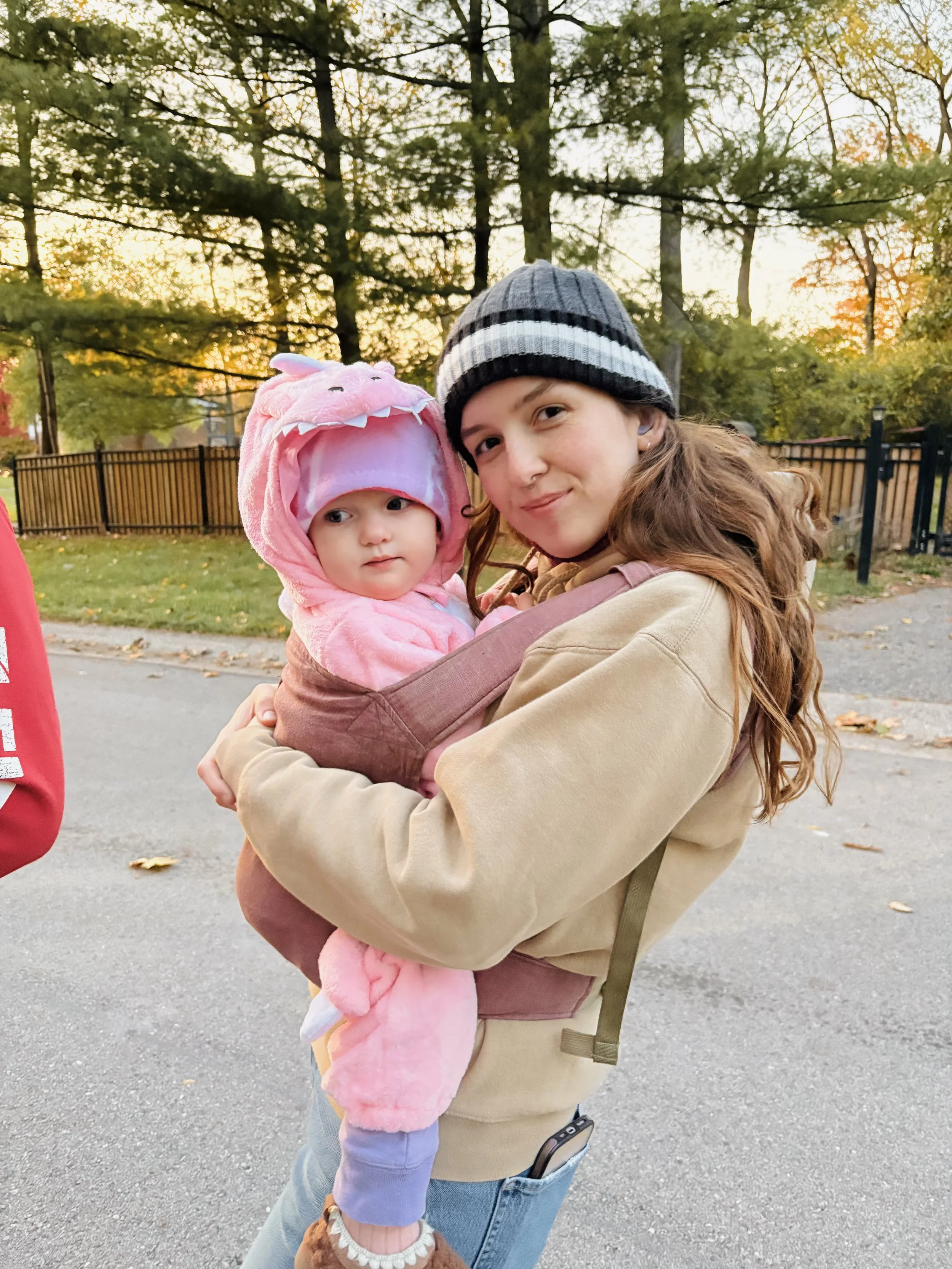A woman with long red hair, wearing a black and gray striped beanie and a beige jacket, holding a young girl dressed in a pink unicorn costume outdoors during sunset.