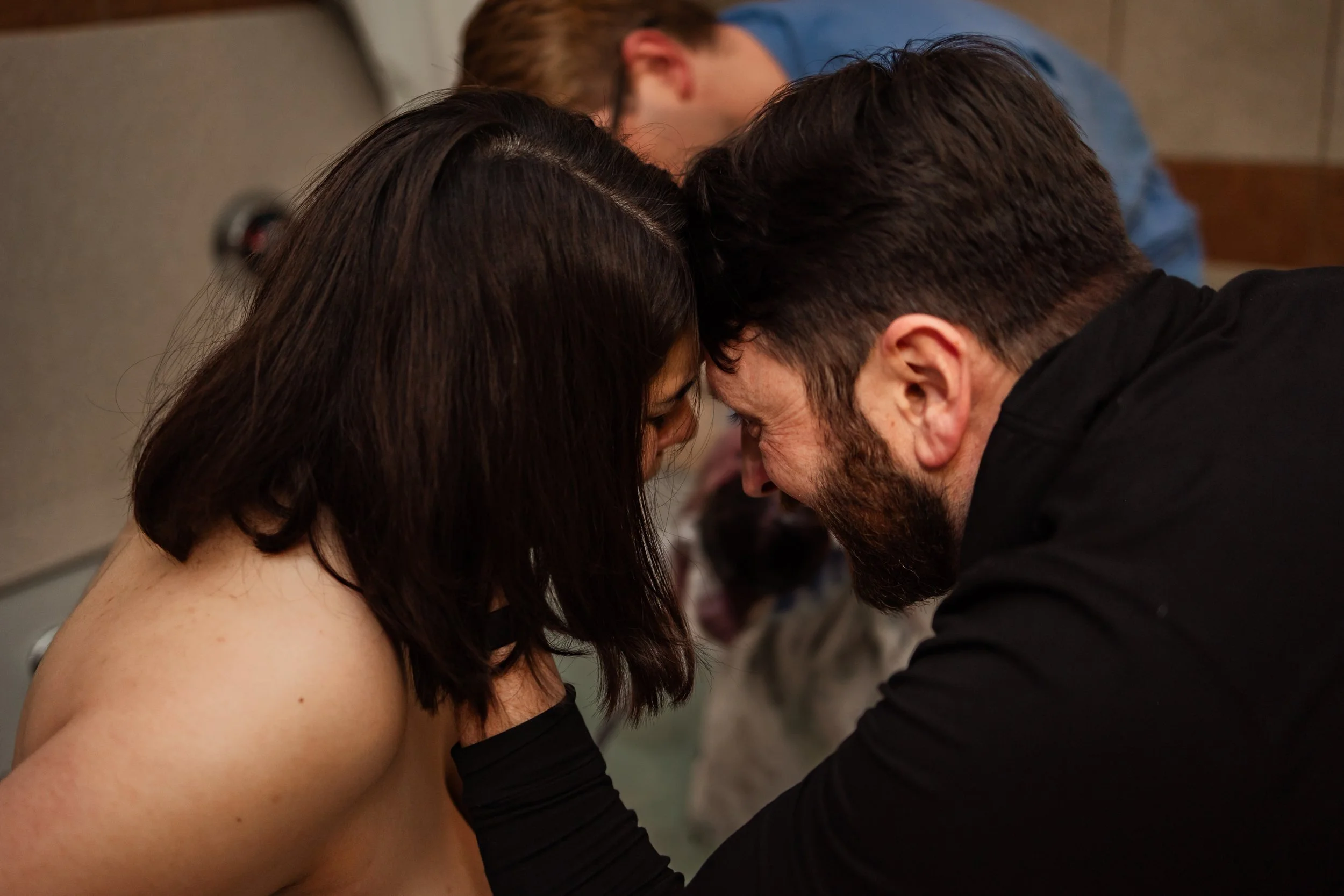 A woman and a man with their foreheads touching indoors at a close-quarters event, with a woman and a dog lying in the background.