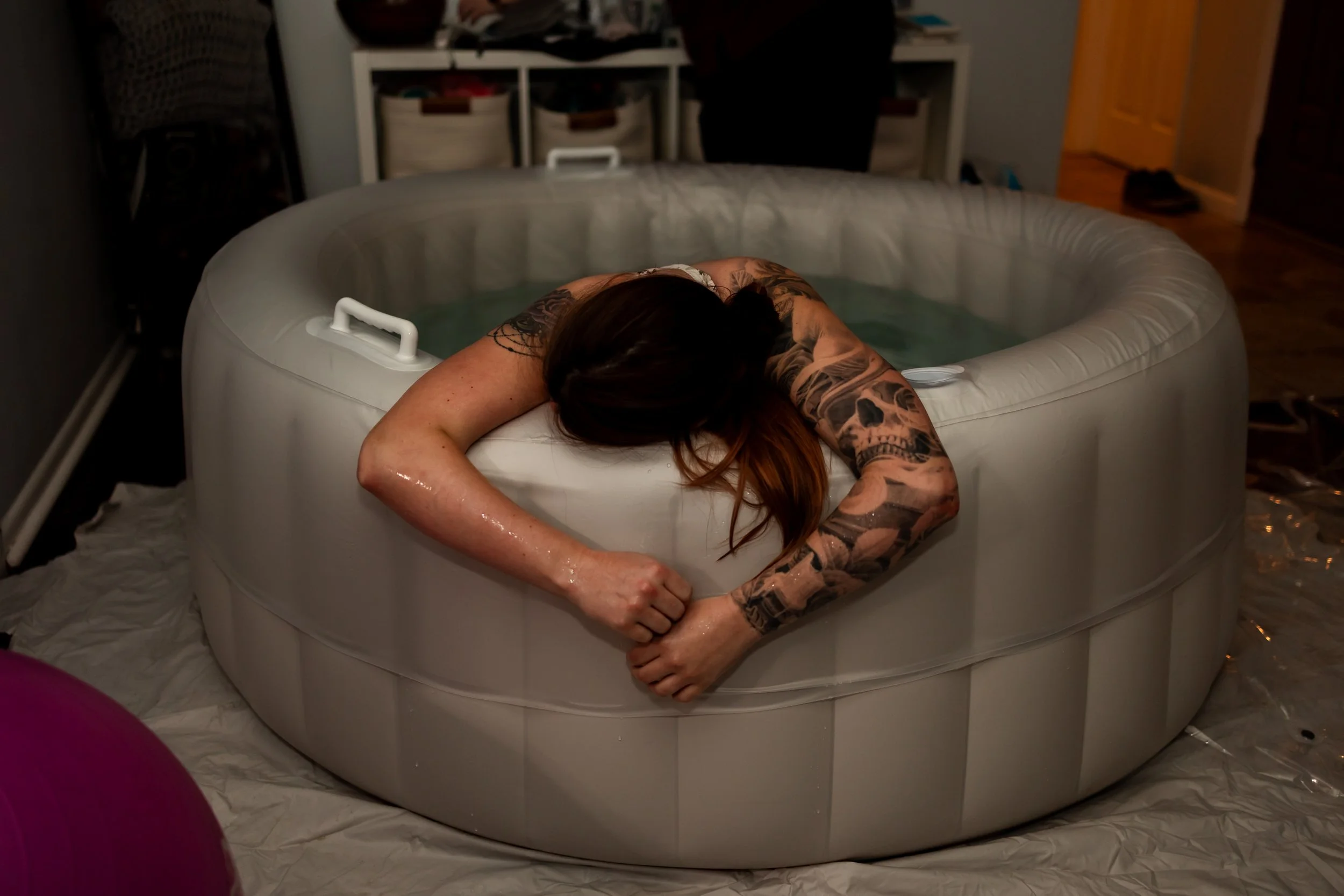 Person with tattoos resting on the edge of an inflatable hot tub filled with water, leaning with arms folded and head down.