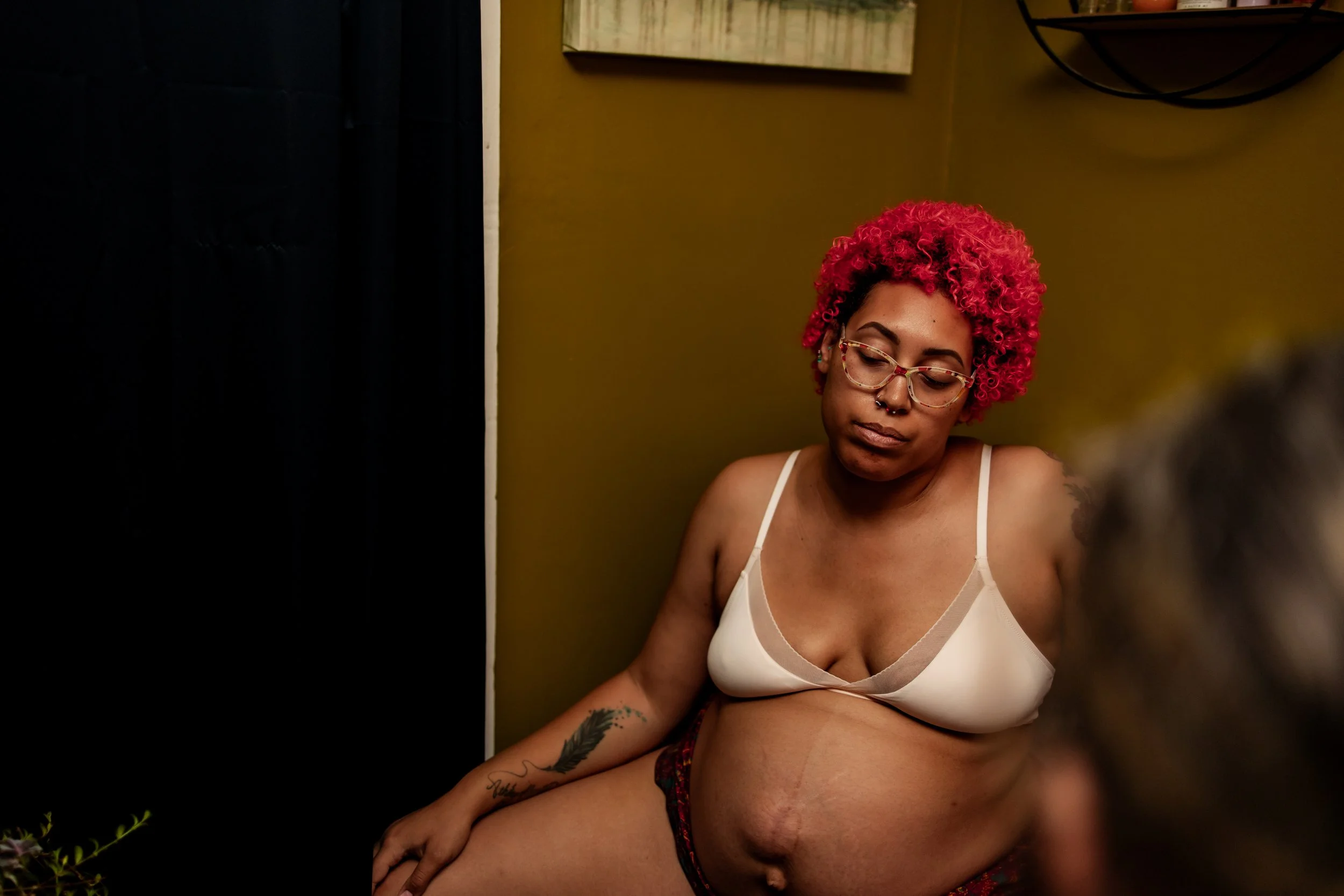A woman with bright pink curly hair, wearing glasses and a white bra, sitting in a room with an olive green wall and a black curtain.
