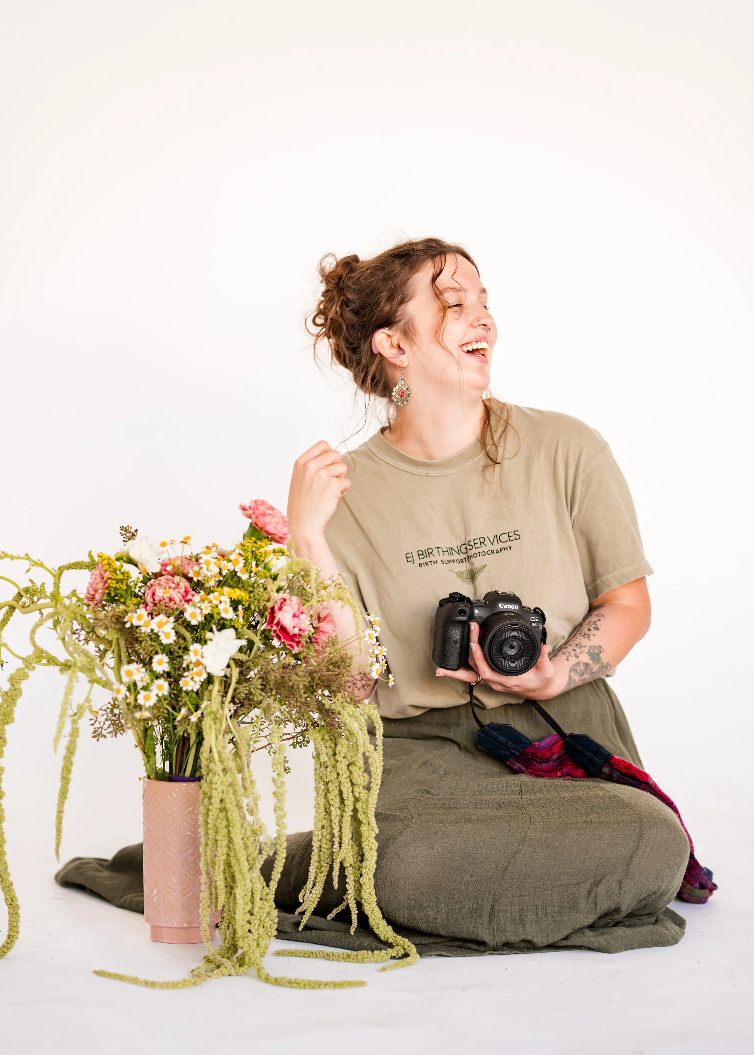 A woman sitting on the floor with her knees up, holding a camera, laughing, next to a large pink vase with a bouquet of flowers. She has curly hair in a bun, wearing earrings, a beige t-shirt with green text, and a long skirt.