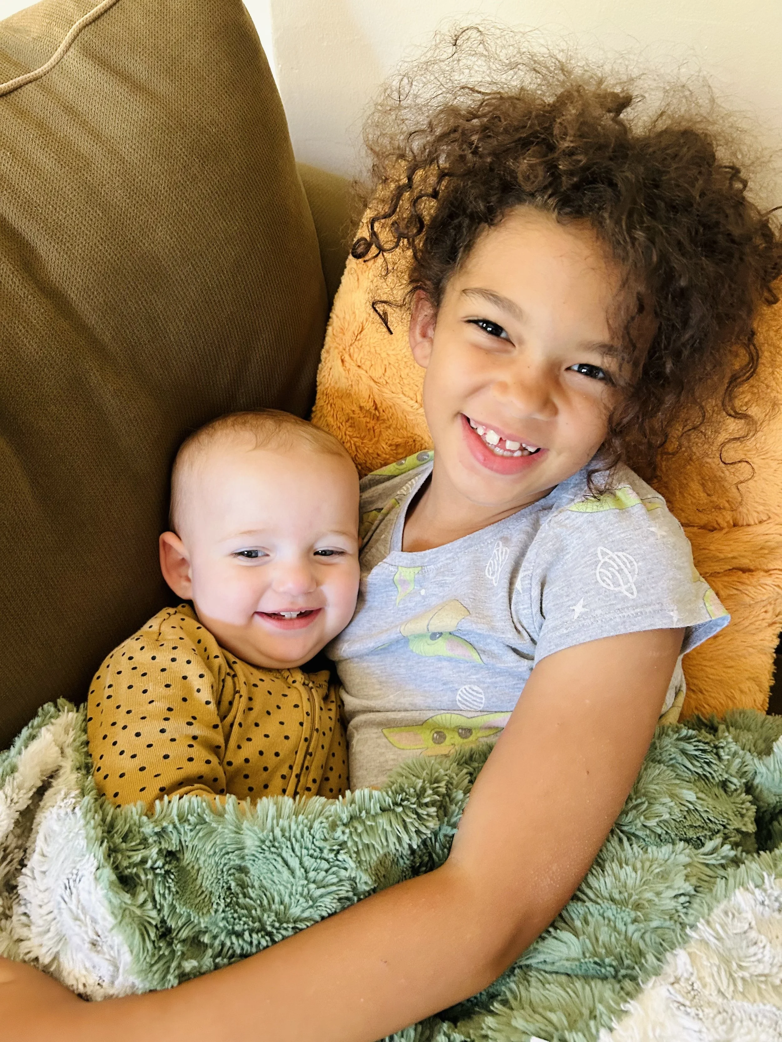 Two young children cuddling and smiling on a couch, wrapped in a green fuzzy blanket. The older girl has curly hair and a gray shirt with cartoon characters, the younger girl has light hair and a yellow polka-dot shirt.