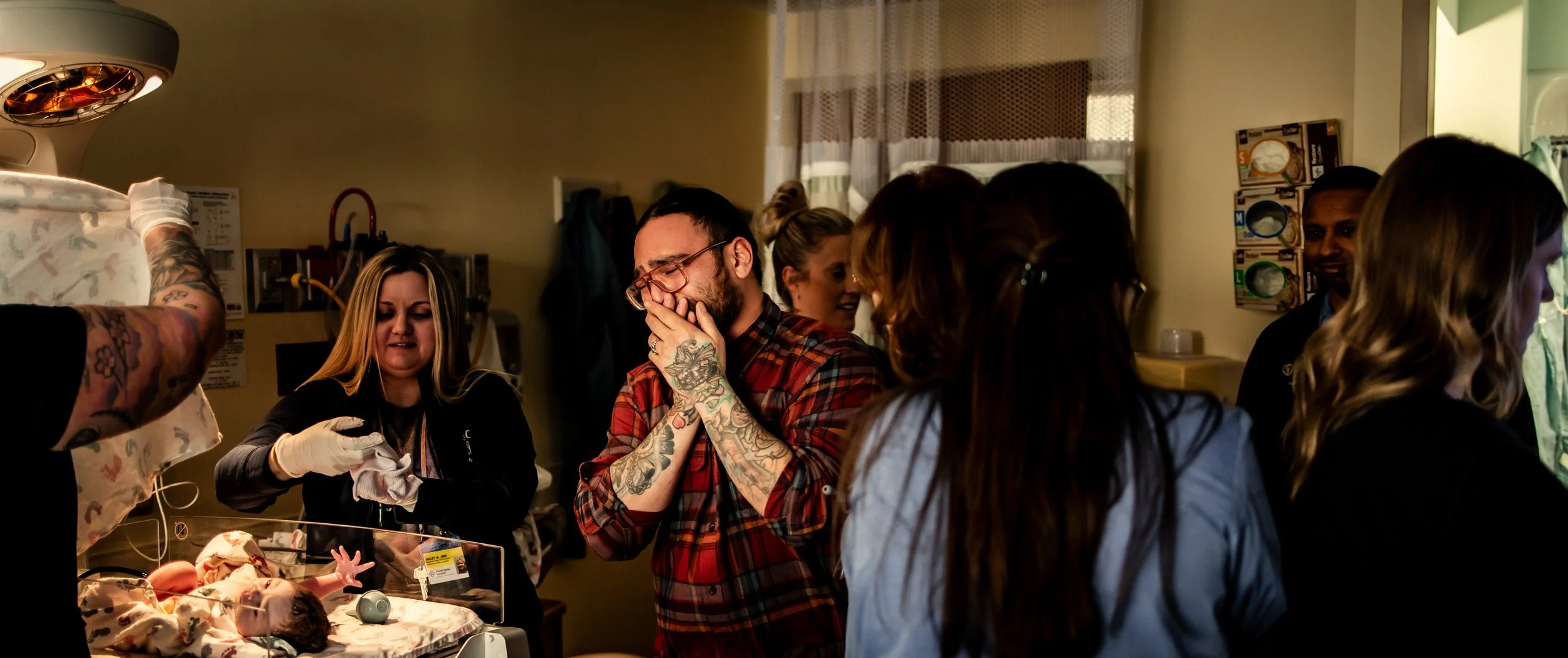 Group of people, including a man with tattoos and glasses, reacting emotionally around a newborn baby in a hospital room. The man is smiling with his hand covering his mouth, and a woman nearby is also smiling. The scene suggests a joyful or emotiona