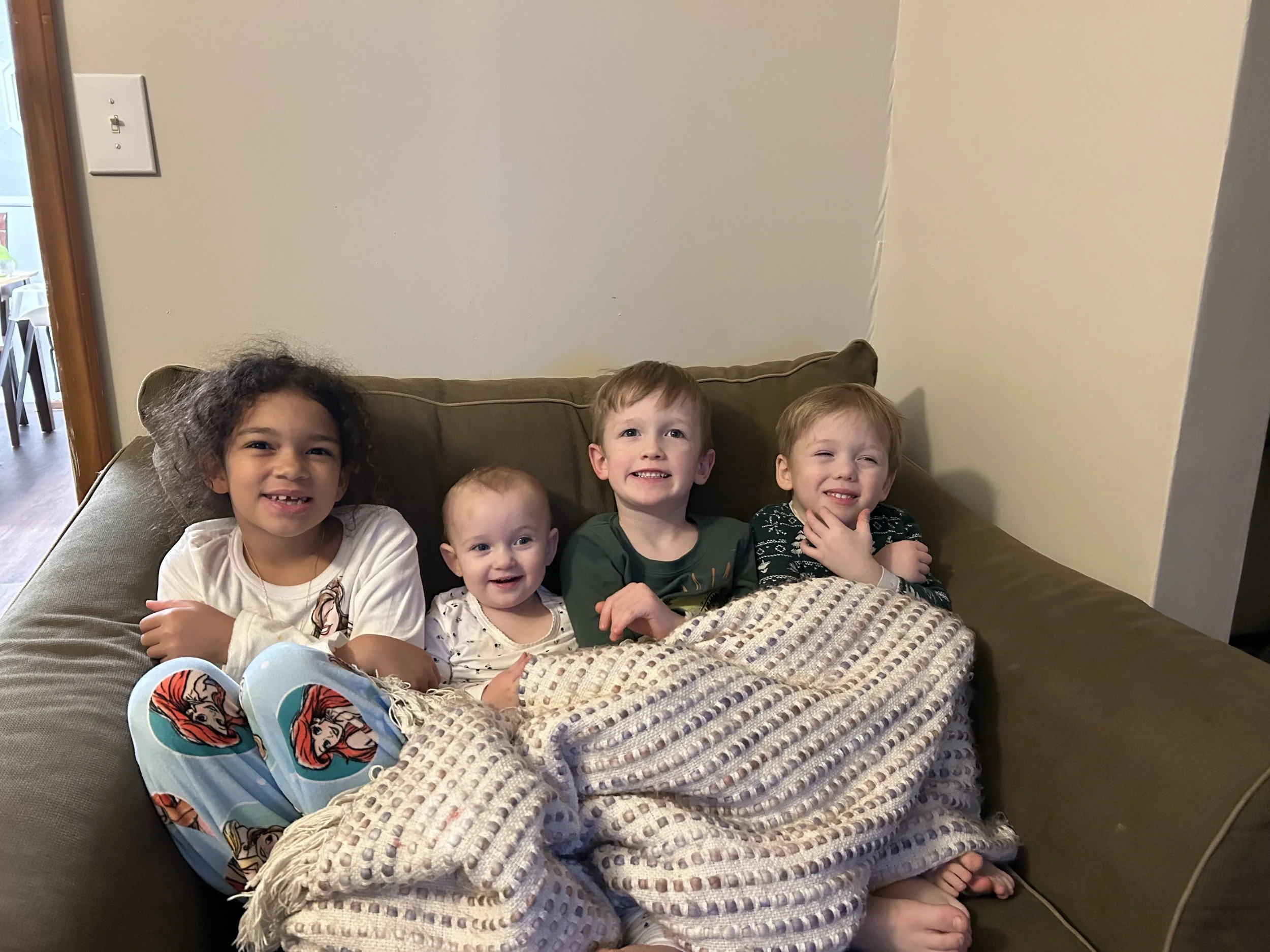 Five children sitting together on a brown couch, smiling and posing for the photo, with a blanket covering their legs.