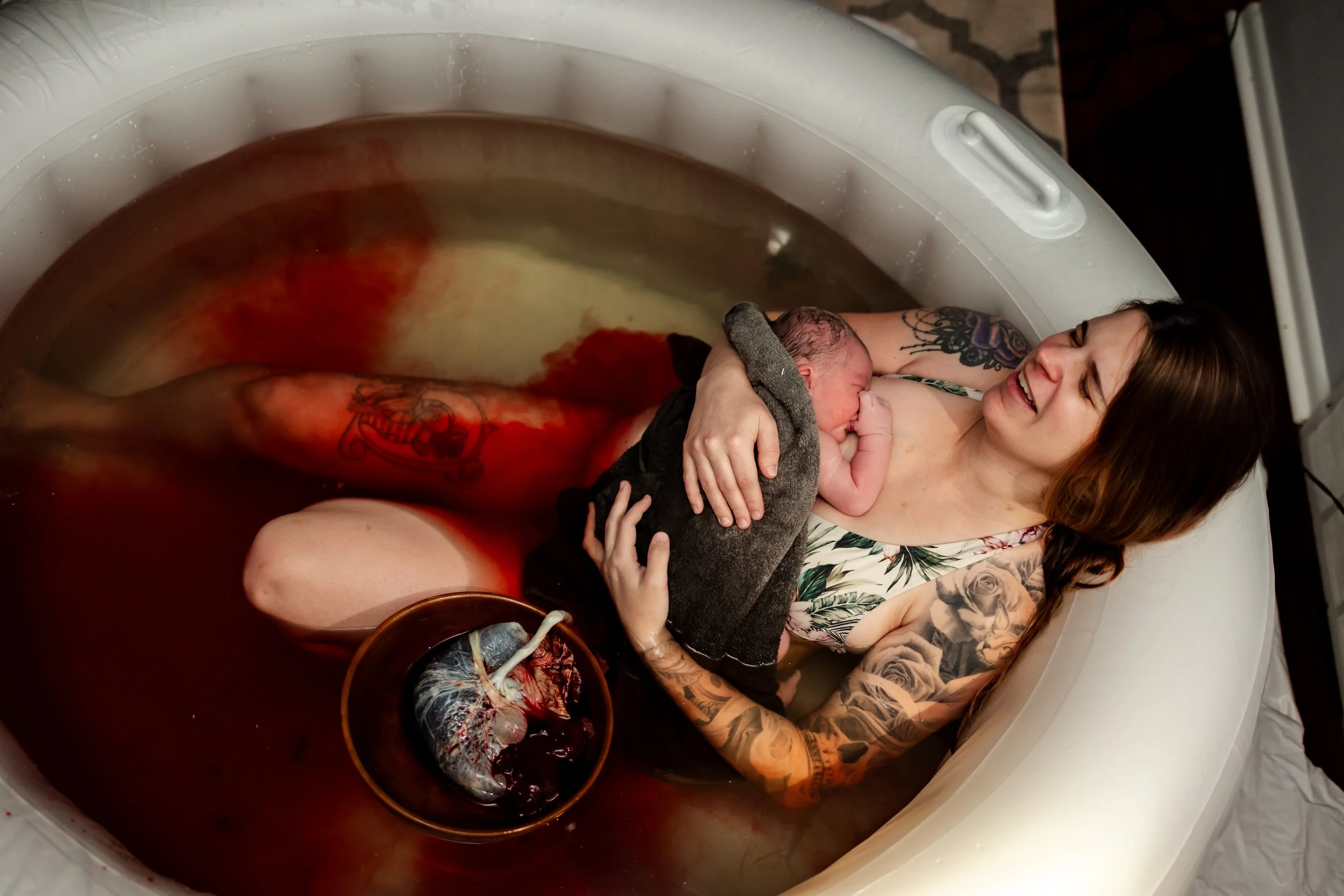 A woman with tattoos cradles a newborn baby in a birthing tub filled with water. The woman is smiling and the baby is resting against her chest. There is a bowl with a human heart and blood inside it floating in the water.