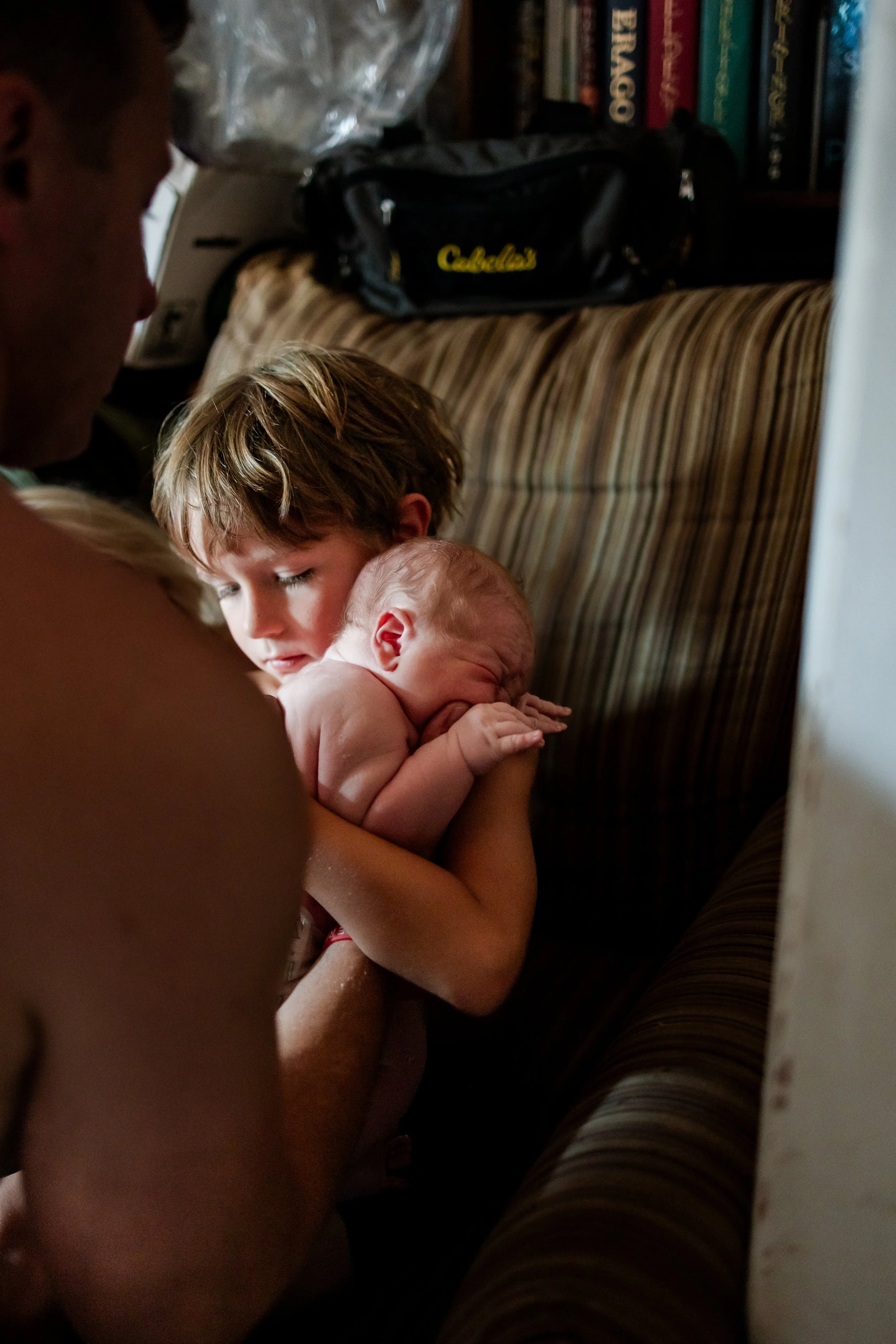 A young boy holds a newborn baby close to a woman, possibly the baby's mother, in a cozy indoor setting.
