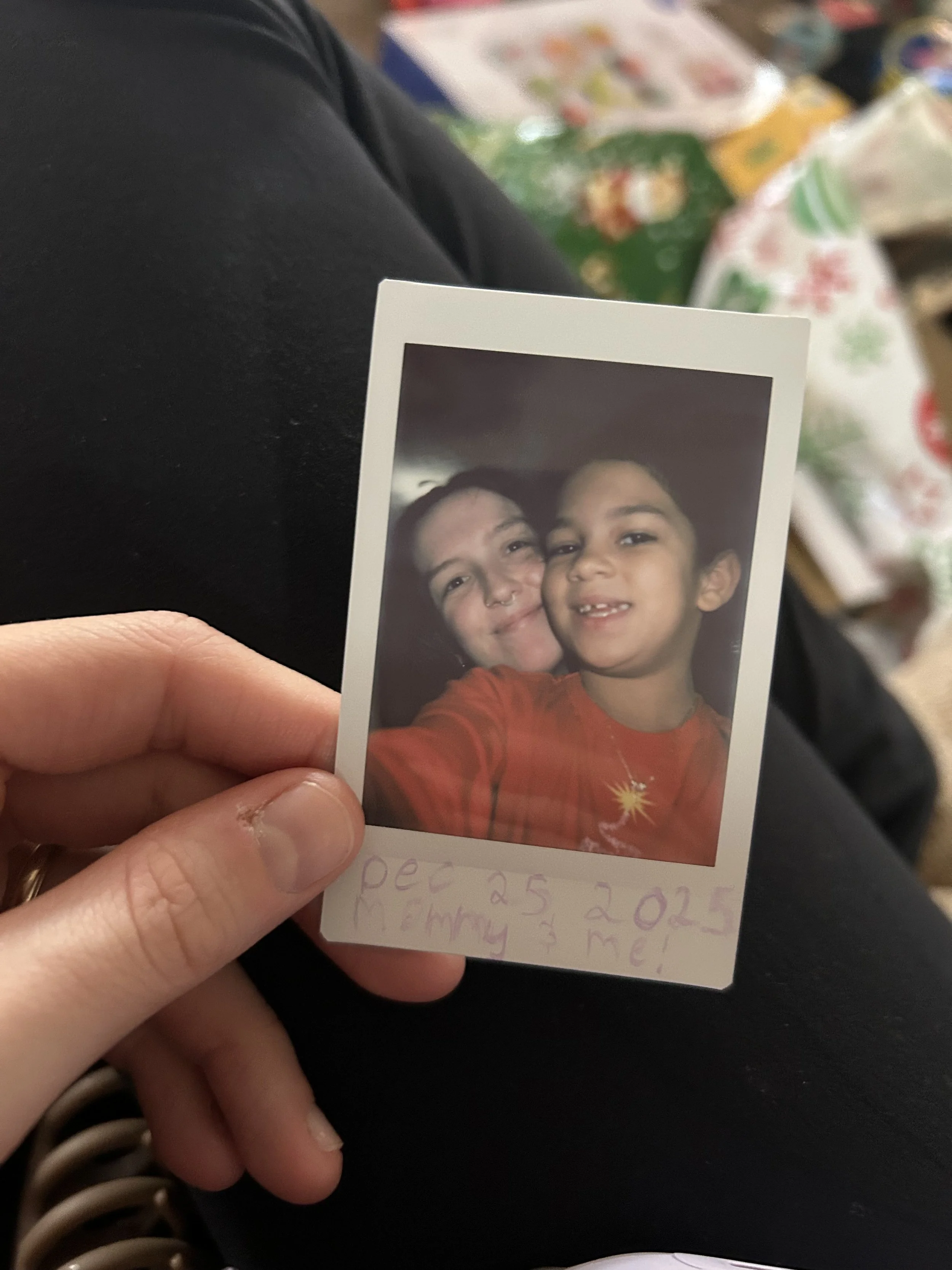 A person holding a selfie photo of two children, one with dark hair and the other with light-colored hair, both smiling and close together, with holiday-themed background objects in the blurry background.