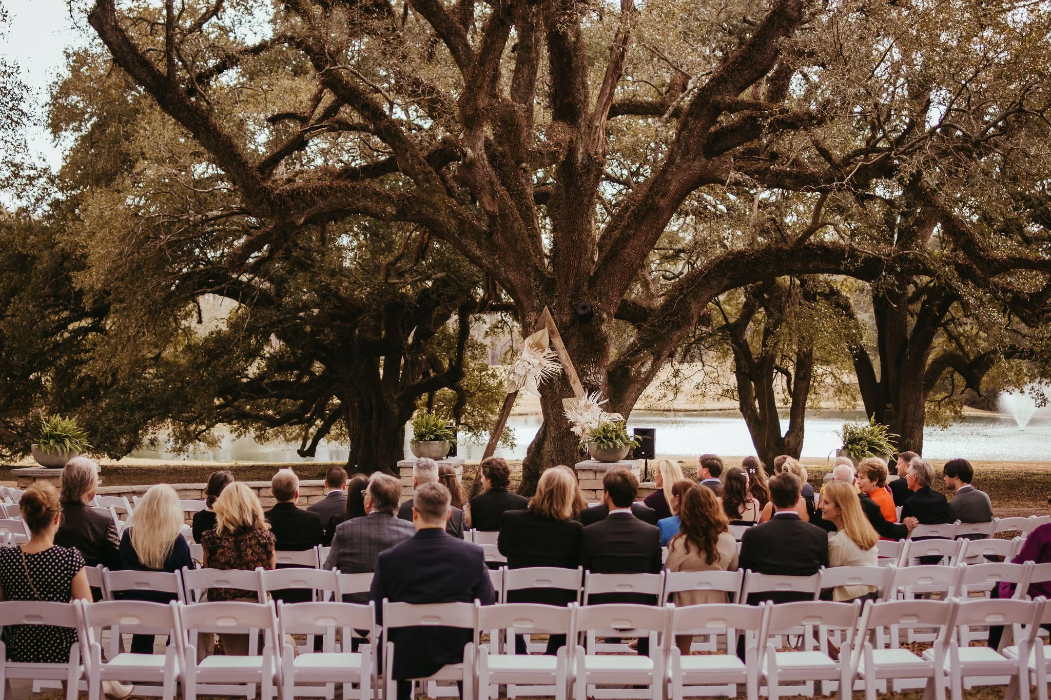 A wedding ceremony taking place outdoors under large oak trees next to a body of water, with guests seated in white chairs facing an arch decorated with flowers.