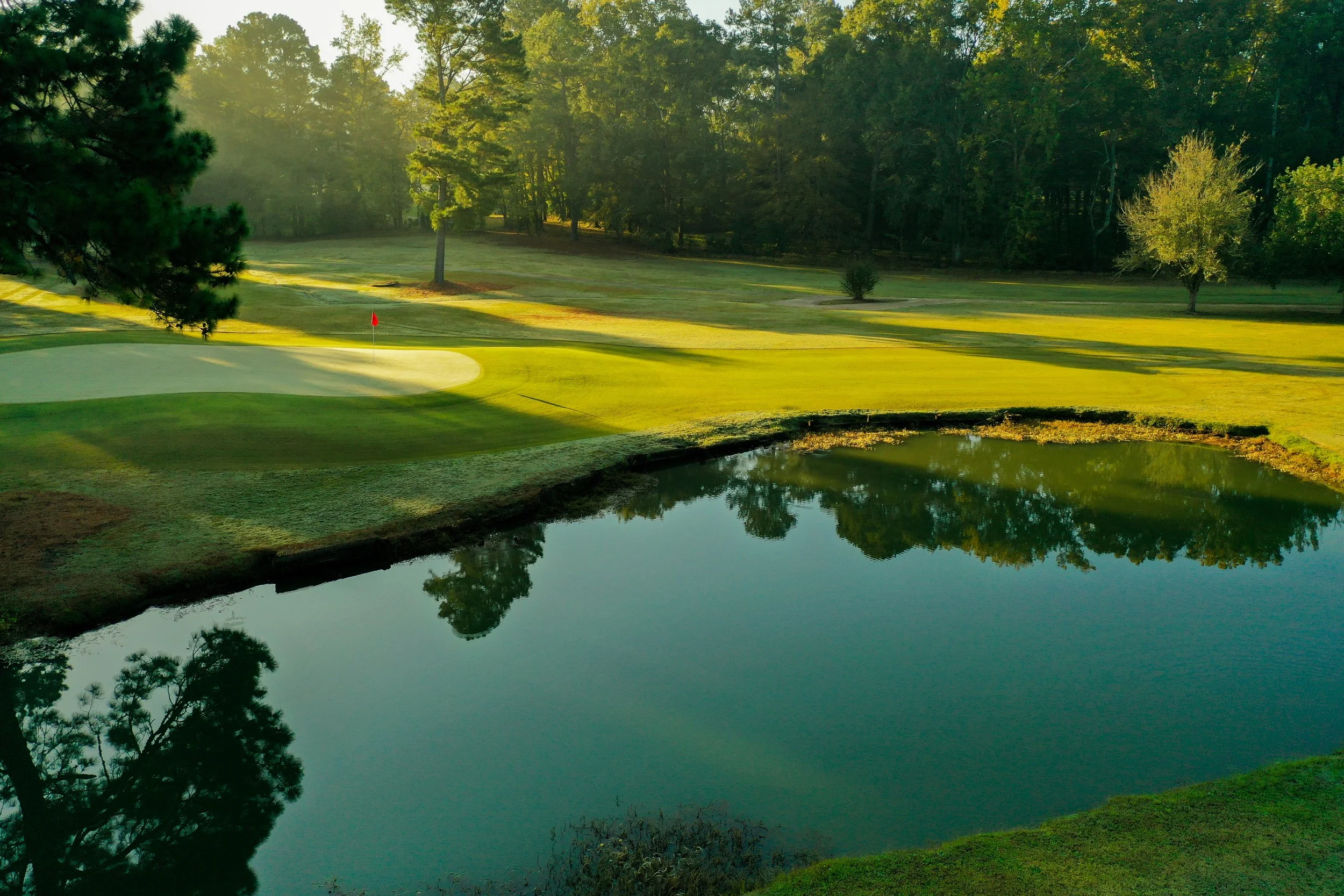 Sunlit golf course with a putting green, a hole flag, and a small pond reflecting surrounding trees and grass.