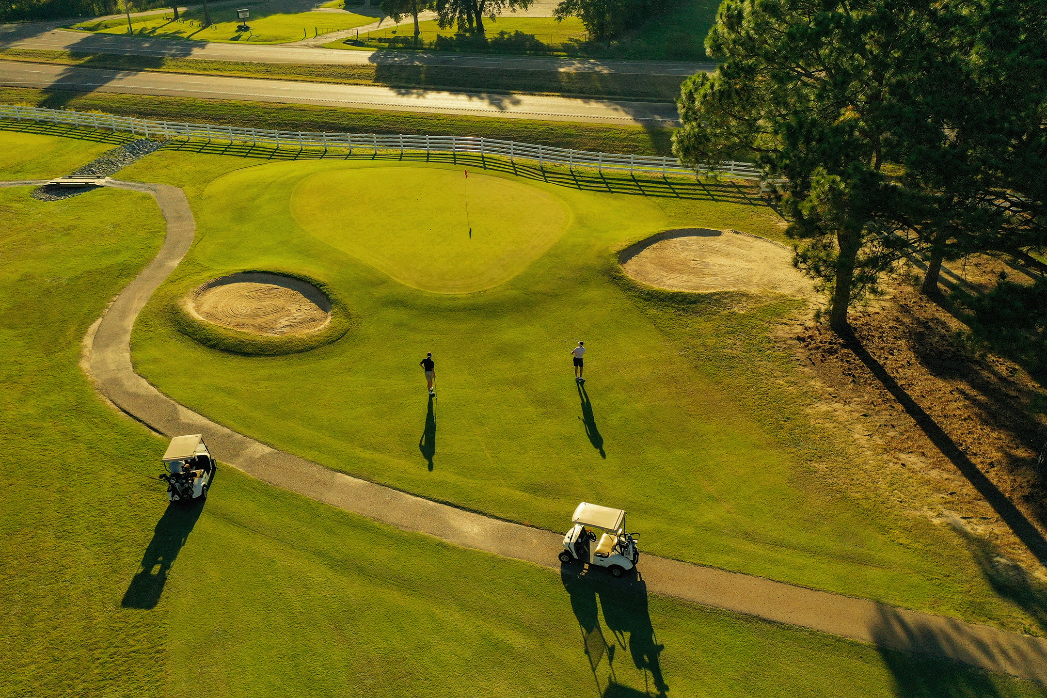 An aerial view of a golf course showing a green with a flag, surrounded by sand bunkers, walking paths, two golf carts, and two golfers playing during late afternoon or early evening.