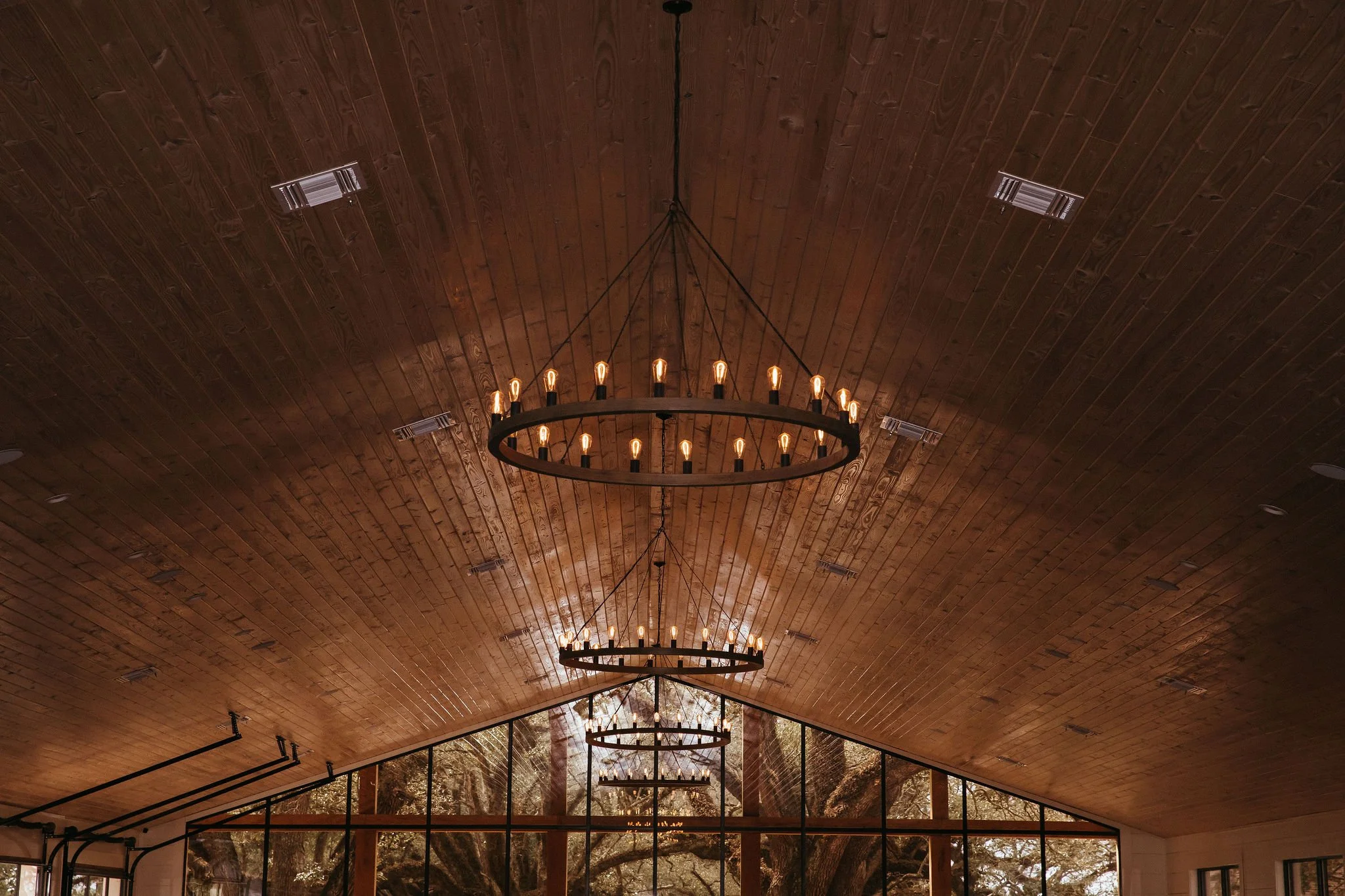 Wood-paneled ceiling with three circular chandeliers hanging, illuminated with multiple light bulbs, large windows showing trees outside.