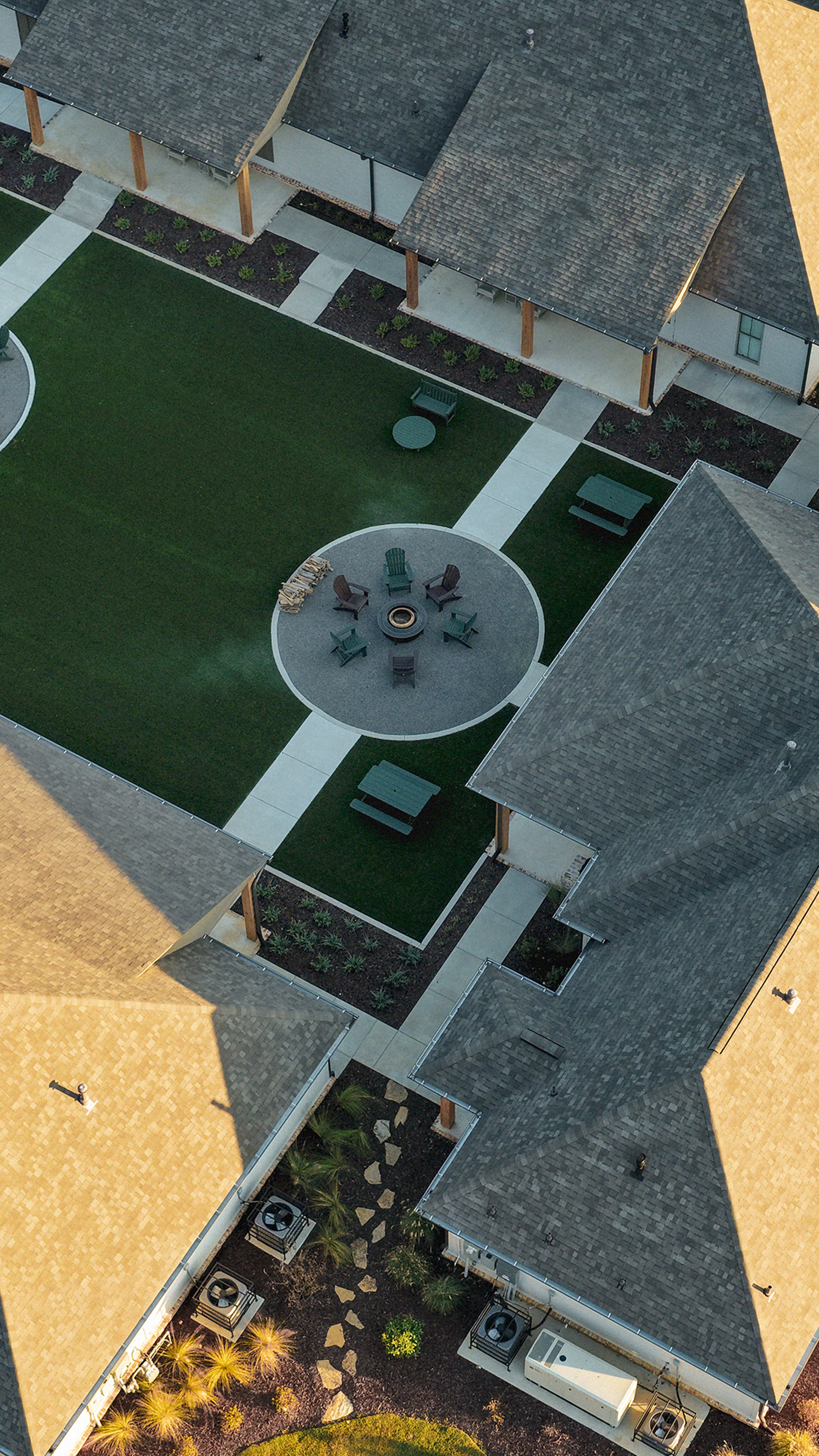 Aerial view of an apartment complex courtyard with a central fire pit area surrounded by chairs, grassy lawns, pathways, benches, plants, and rooftops of the surrounding buildings.