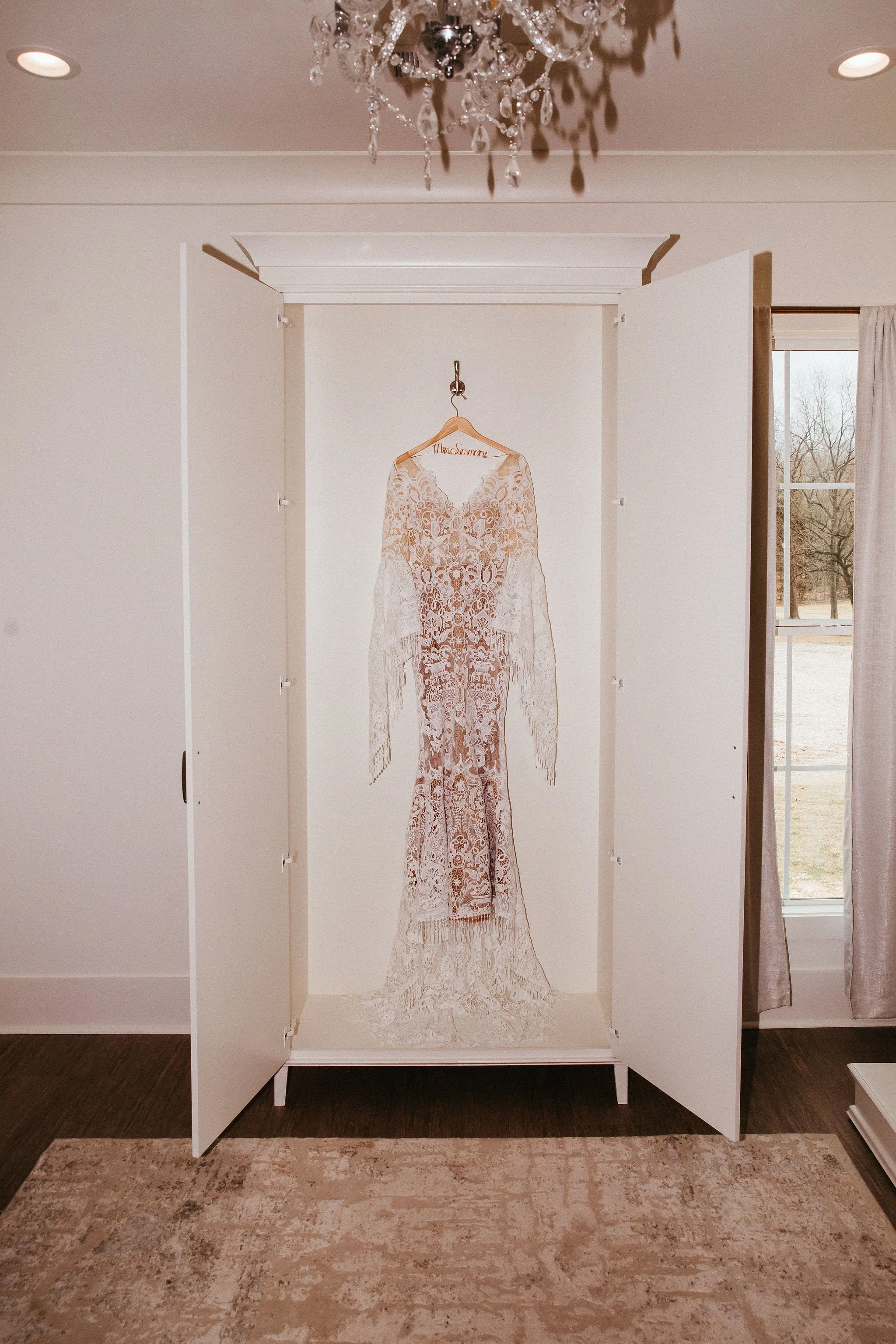 A lacy wedding dress hanging inside an open white wardrobe, with a chandelier above and a window with curtains to the right, showing an outdoor scene with trees.