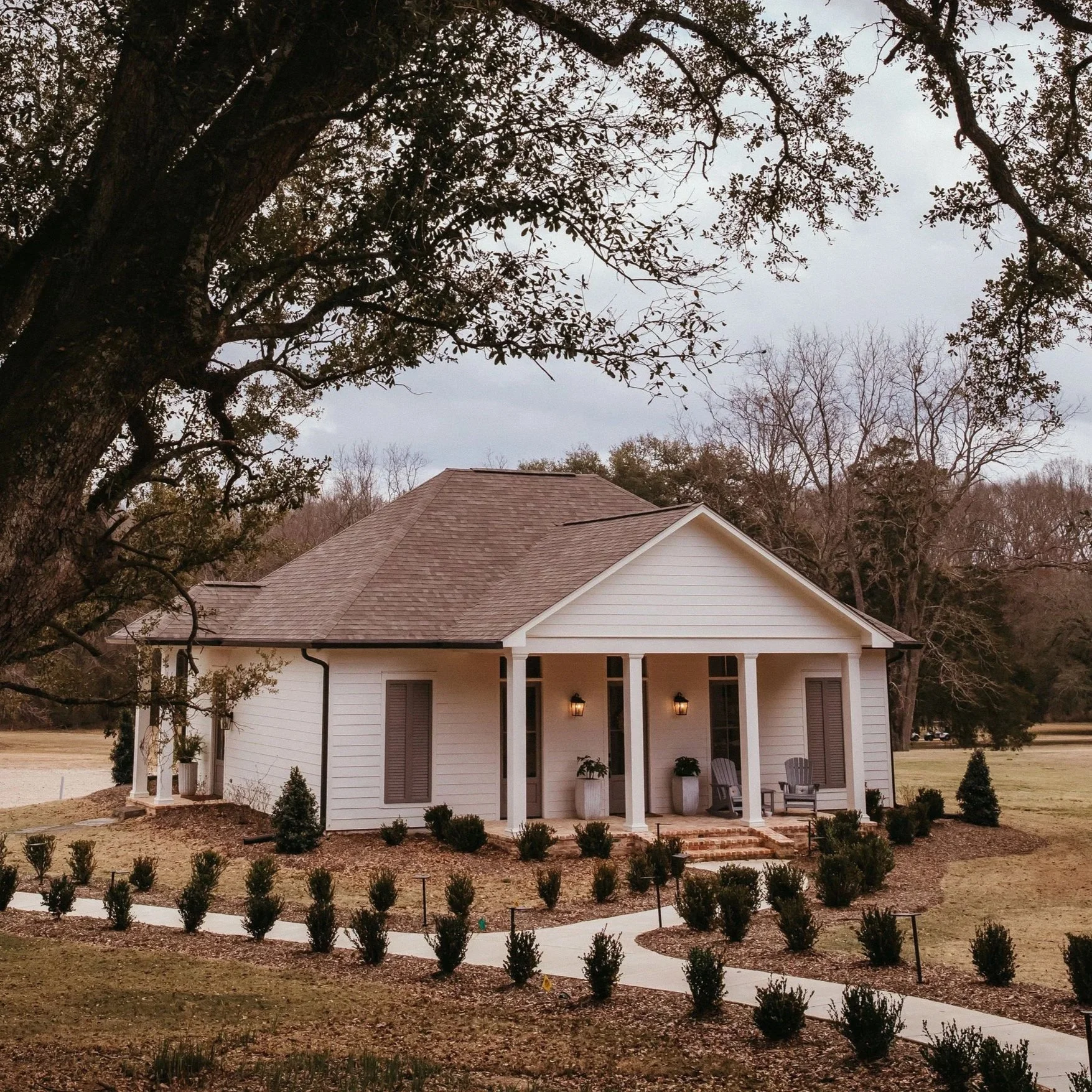 A white house with a front porch, two chairs, potted plants, and outdoor lighting, surrounded by a manicured lawn with small bushes and trees in the background.