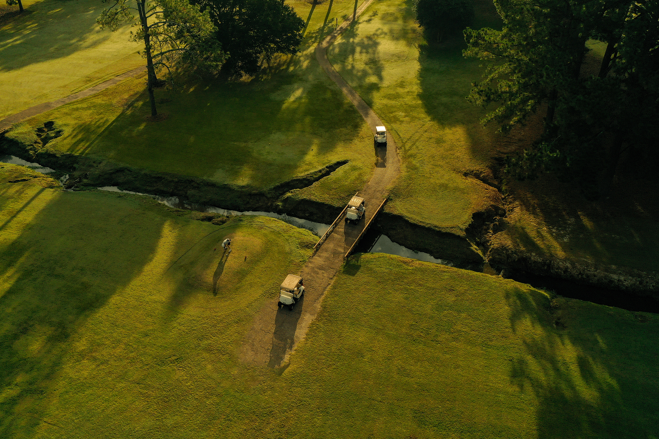 An aerial view of a golf course with a small wooden bridge crossing a narrow stream. Three golf carts are on the grassy path near the bridge, and one golfer is putting on the green. Tall trees cast shadows across the area of the fairway.