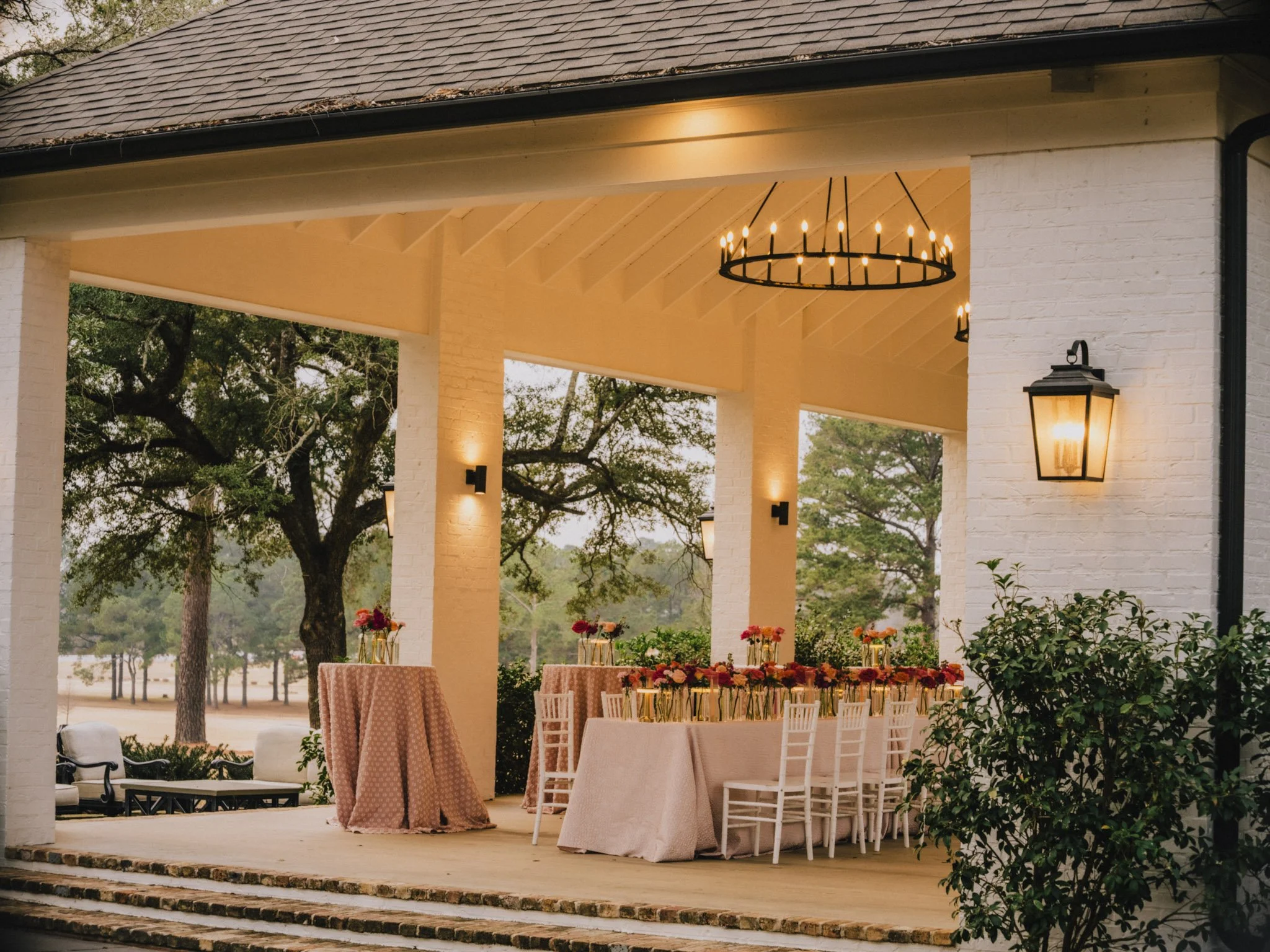 Outdoor event space dining area with a rectangular table decorated with flowers, surrounded by white chairs, under a covered porch with hanging chandeliers and wall lanterns, in a green pavilion outdoor setting.