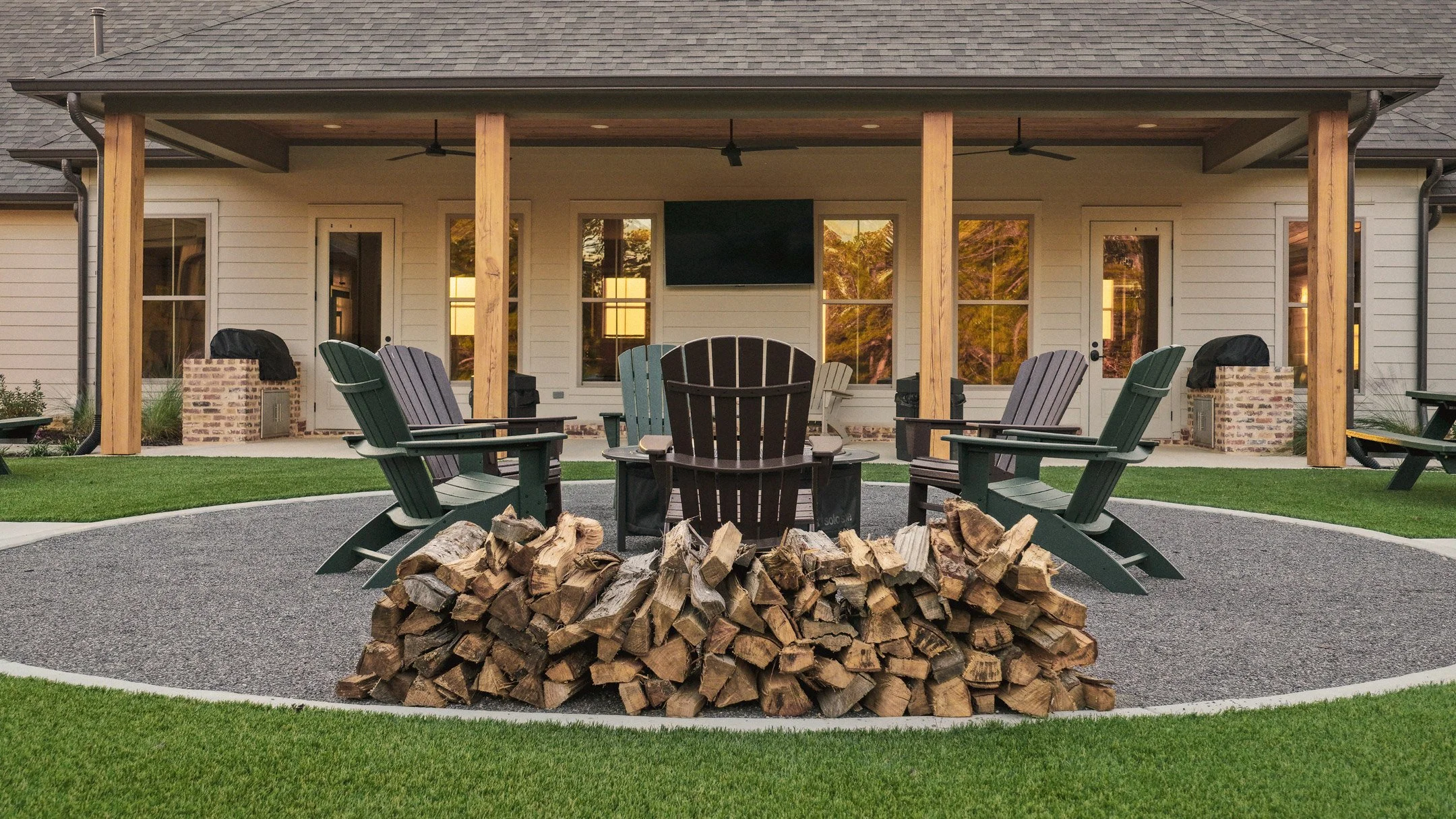Outdoor fire pit and covered porch at the Bunk House group lodging facility at Live Oaks Golf Club in Jackson, Mississippi, featuring stacked firewood, green Adirondack chairs arranged in a circle, ceiling fans, an outdoor mounted TV, and warm lights