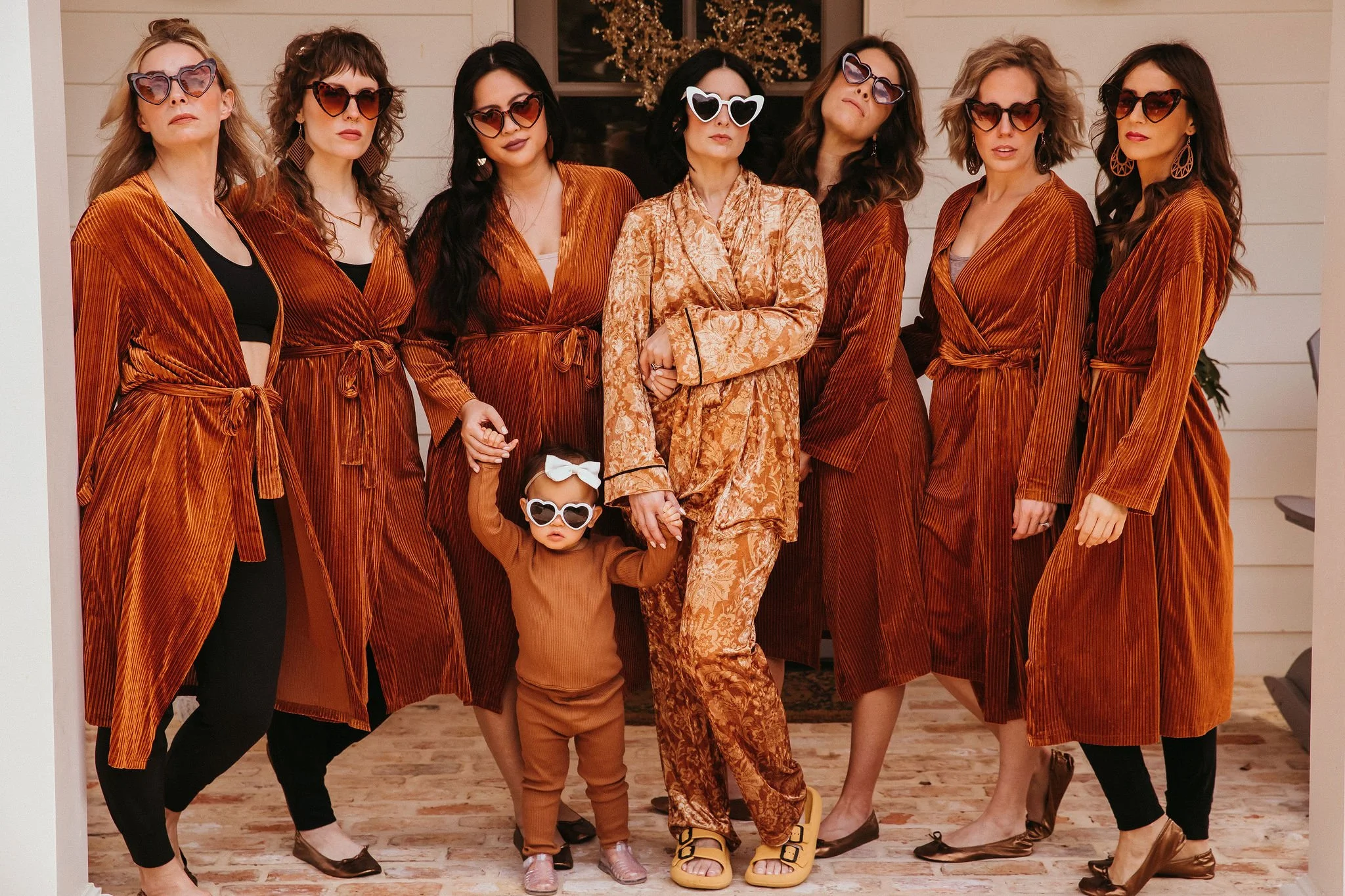 A group of eight women and one young girl dressed in matching rust-colored outfits, wearing sunglasses, posing indoors against a beige wall with a window and holiday decorations, some holding hands.